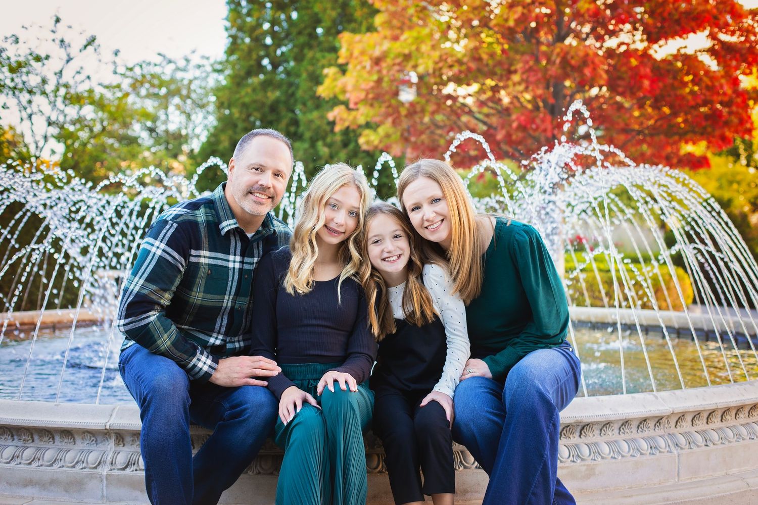 Family of four smiling by a fountain with vibrant red autumn trees behind them.