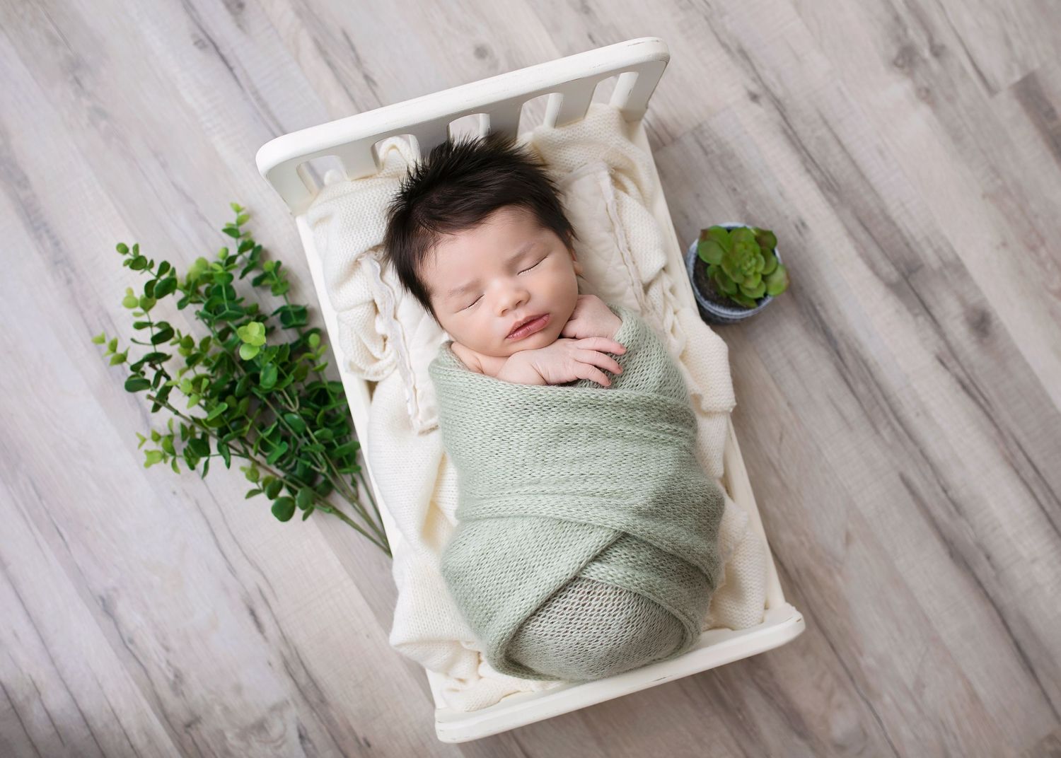 Peaceful newborn baby swaddled in green wrap sleeping in white mini crib surrounded by plants.