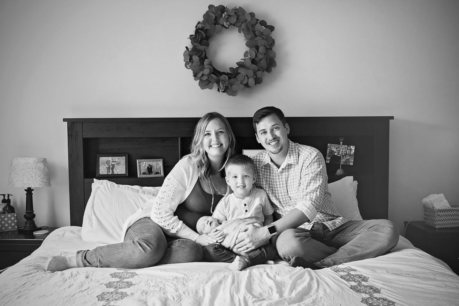 Black and white photo of smiling family of three sitting together on a bed with wreath decor.