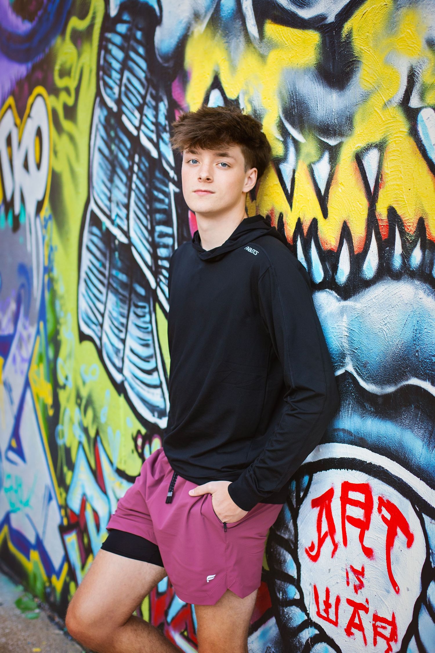 Teen boy in black hoodie and maroon shorts posing in front of colorful graffiti wall.