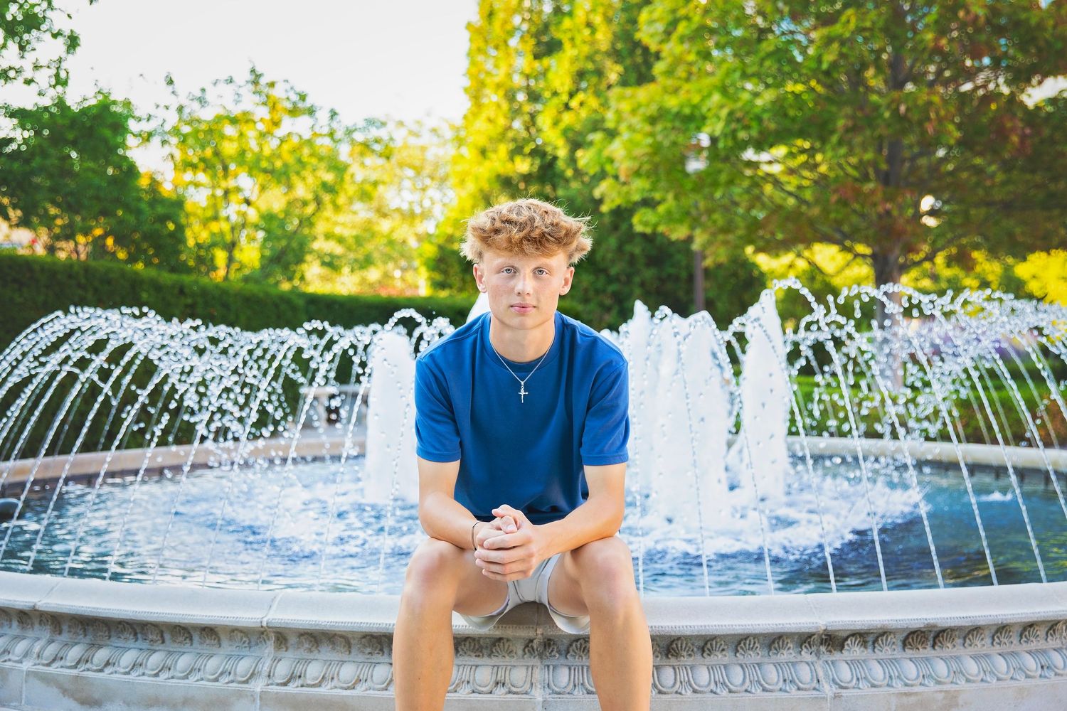 Teen boy with curly hair sits on edge of outdoor water fountain in sunny green park.