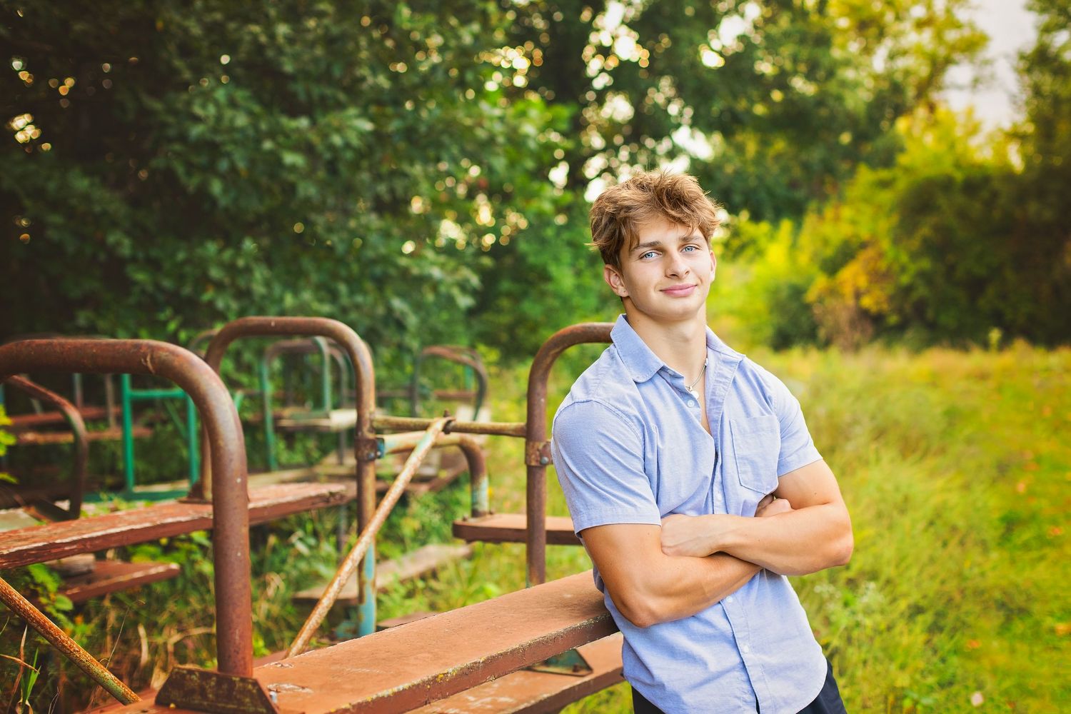 Young man with curly blond hair stands arms crossed near rusty metal equipment in a lush green outdoor setting.