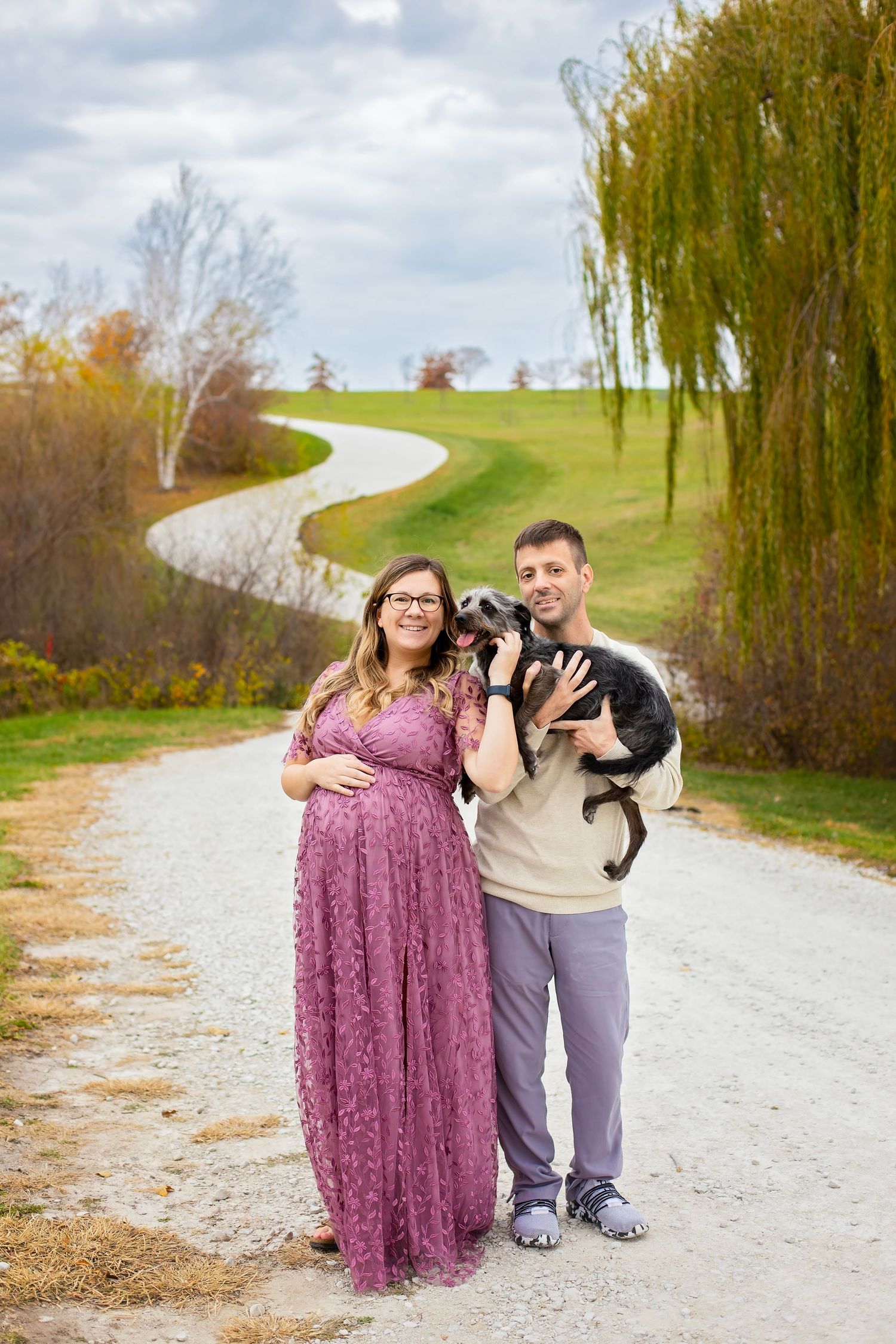 Expectant couple in purple dress and beige sweater hold their dog on a winding country path.