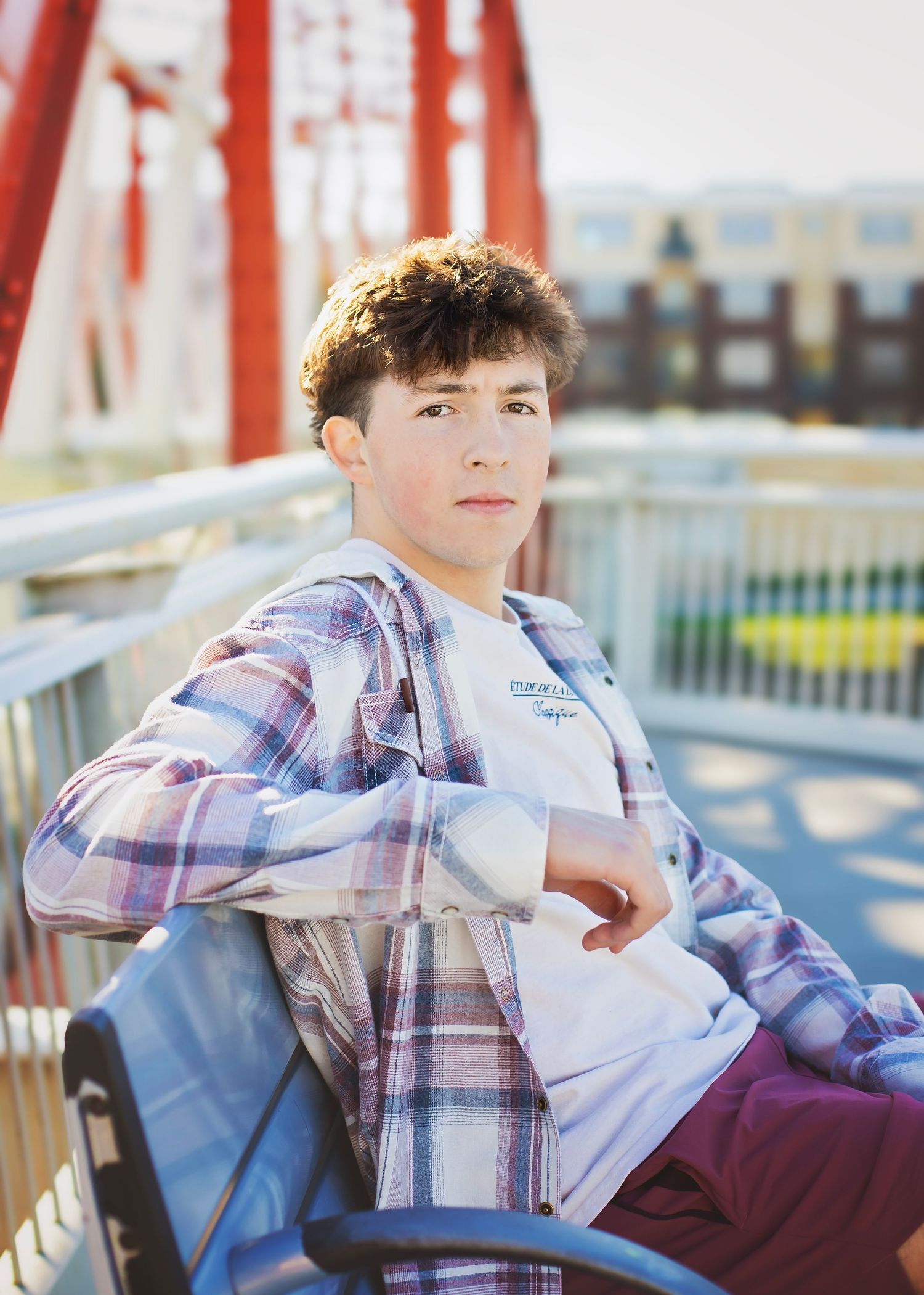 Teen boy in plaid shirt poses on bridge with red steel structure background.