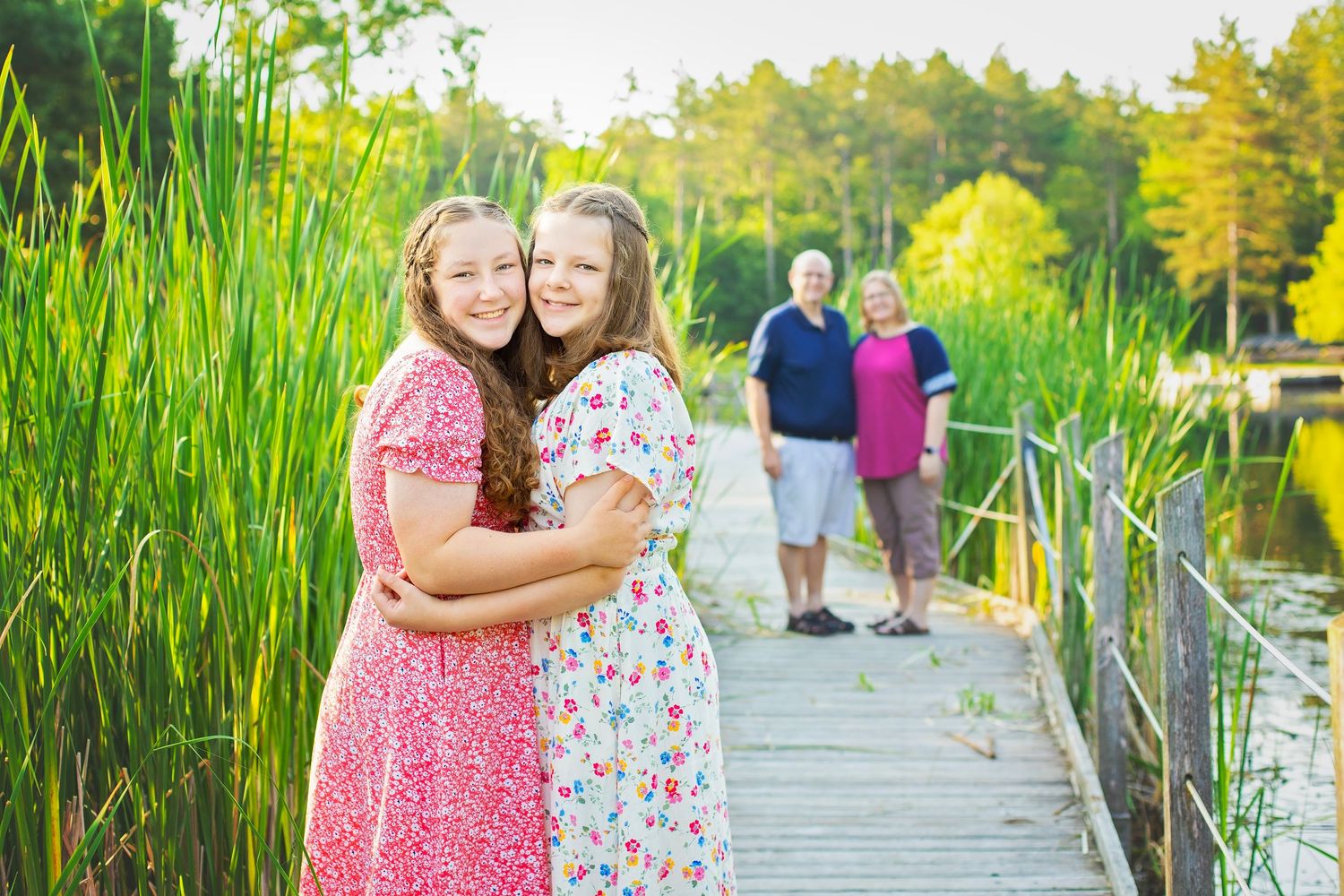 Two girls in matching dresses hugging on wooden dock with grandparents standing behind them.