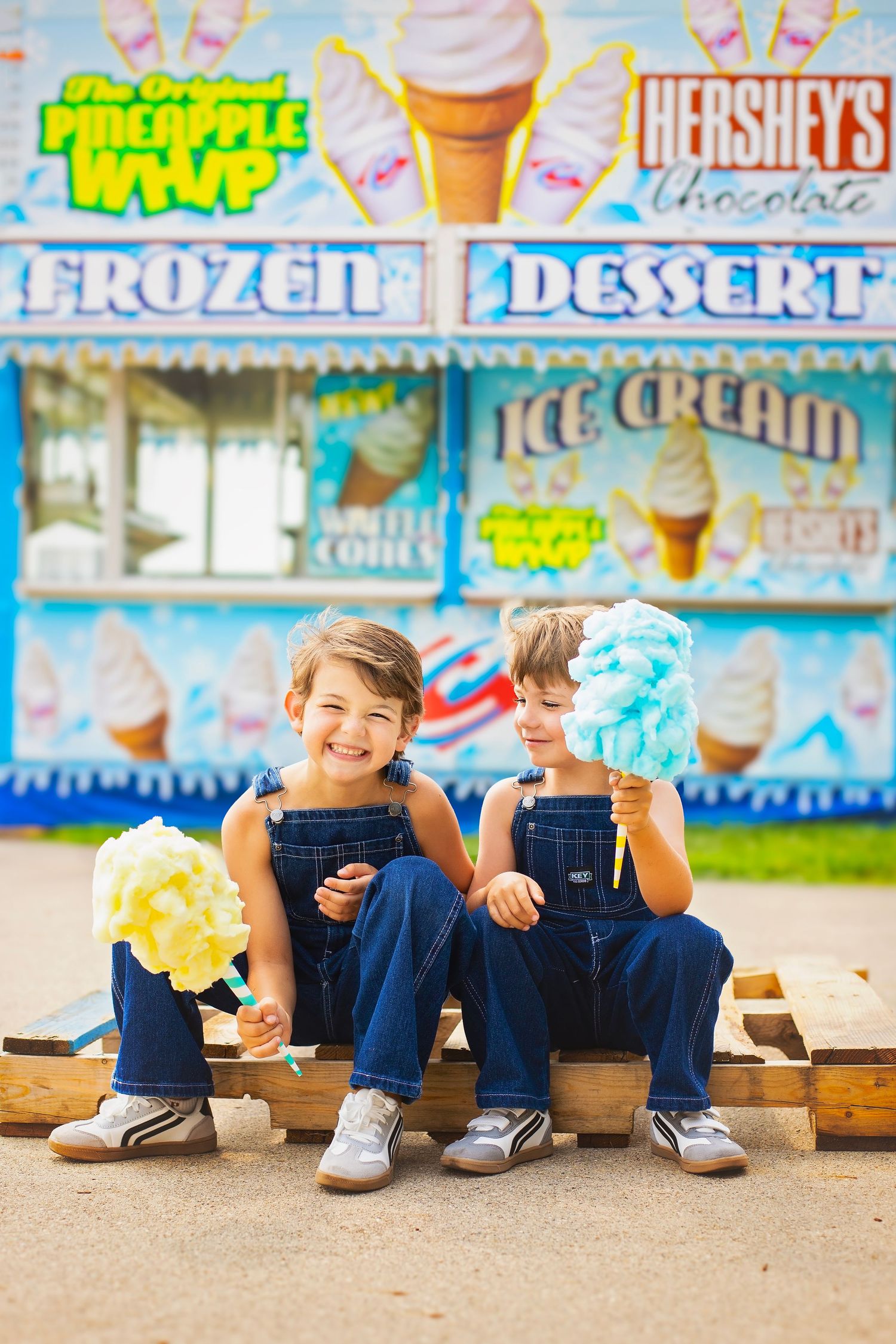 Two young boys in denim overalls enjoy colorful snow cones in front of carnival ice cream stand.