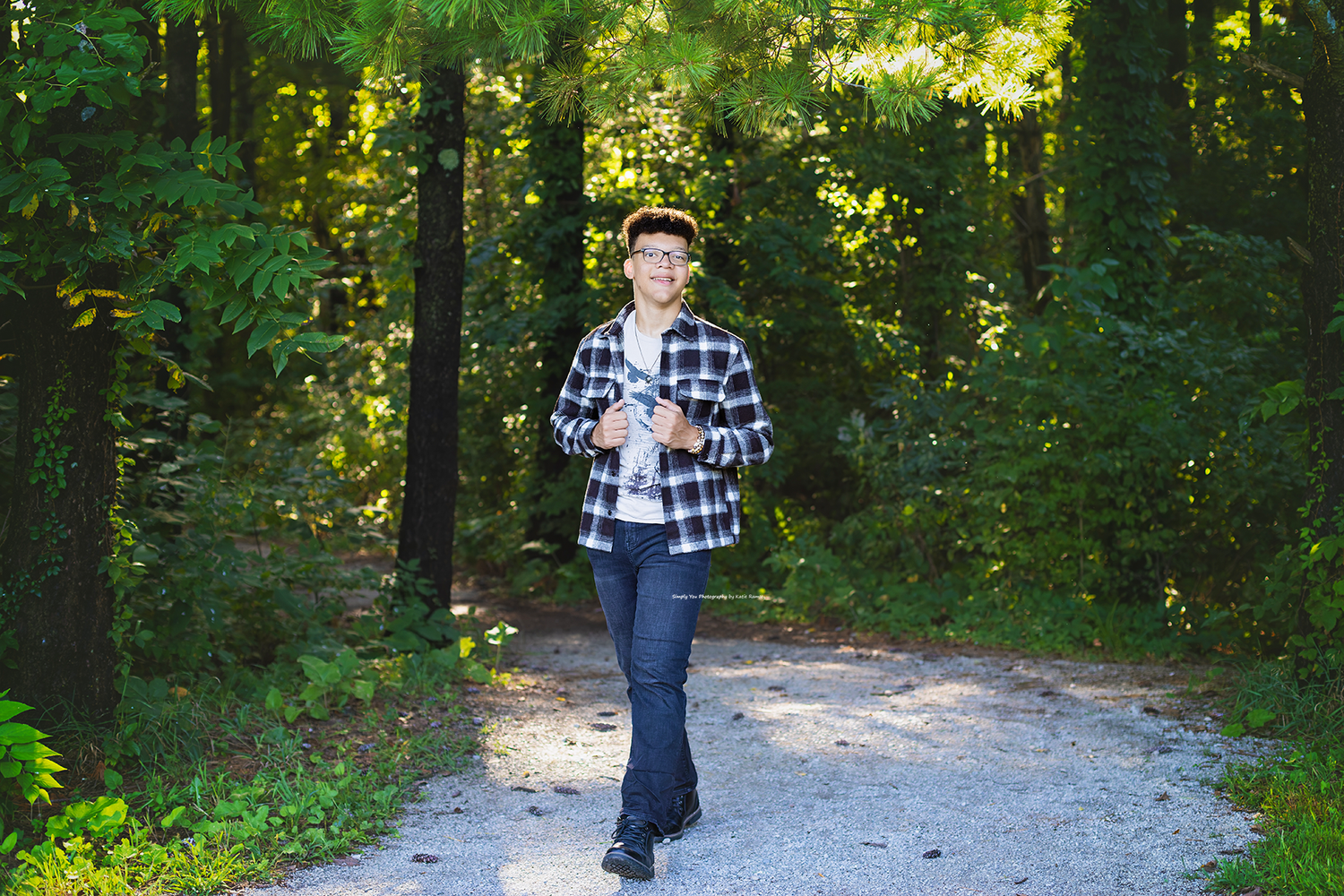 Young teen in plaid shirt and cap walking on forest path surrounded by green trees.