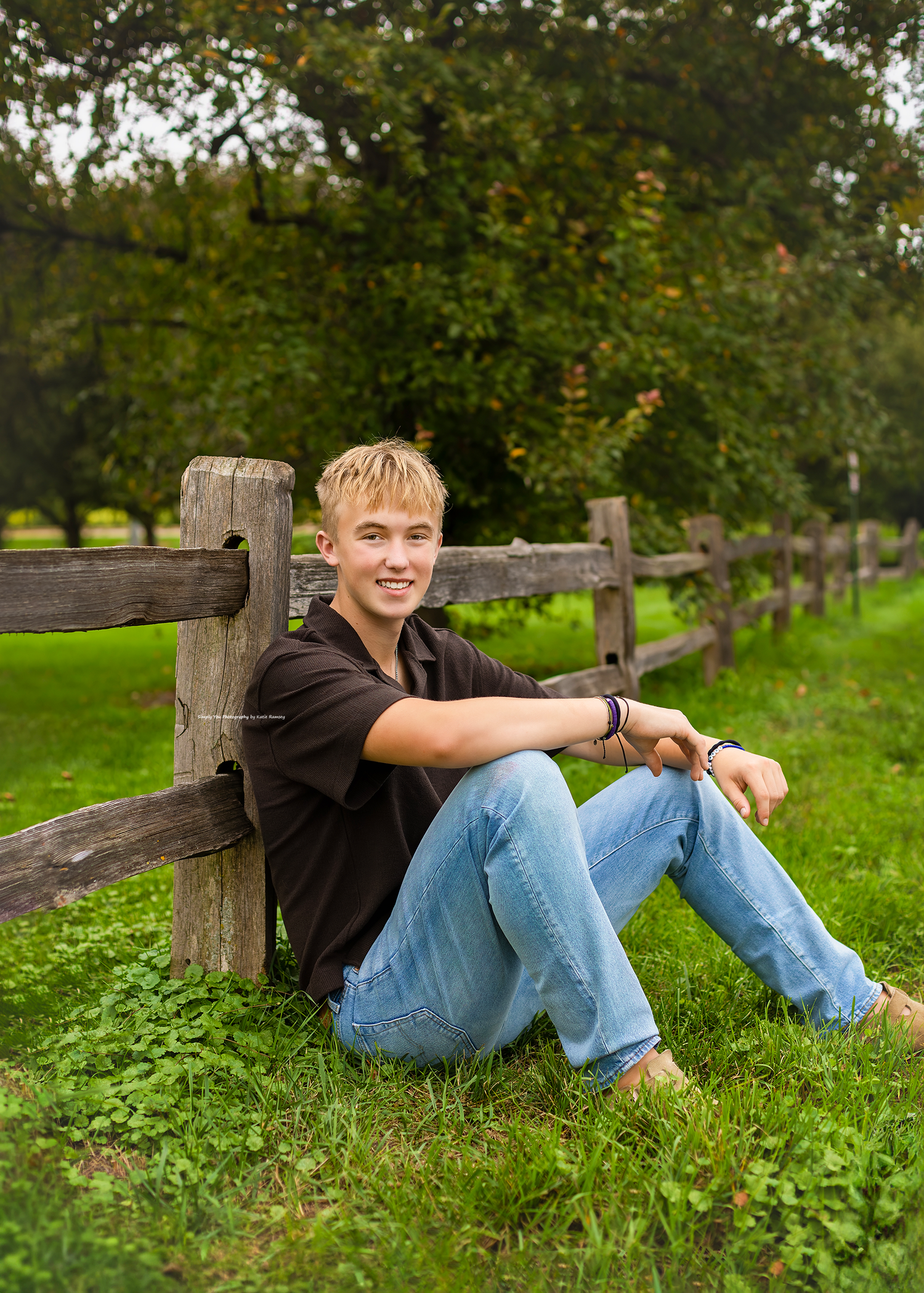 Teen boy in jeans and black shirt sits against rustic wooden fence on green grass with trees in background.