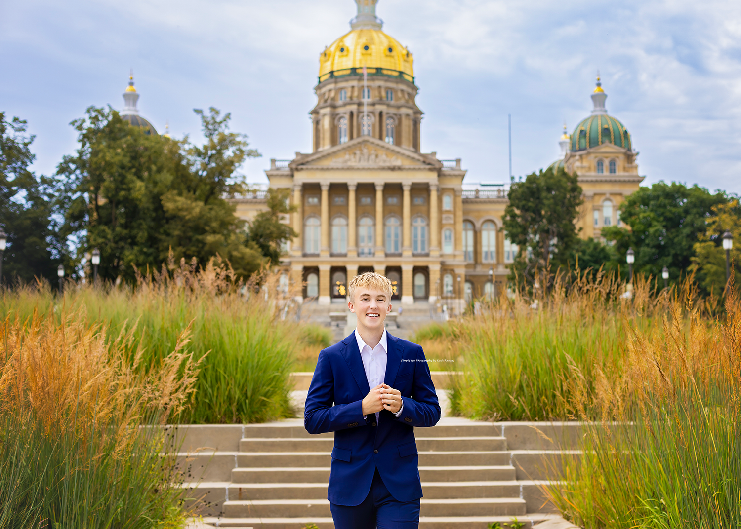 Young man in blue suit stands before Iowa State Capitol building with golden dome and ornamental grasses.