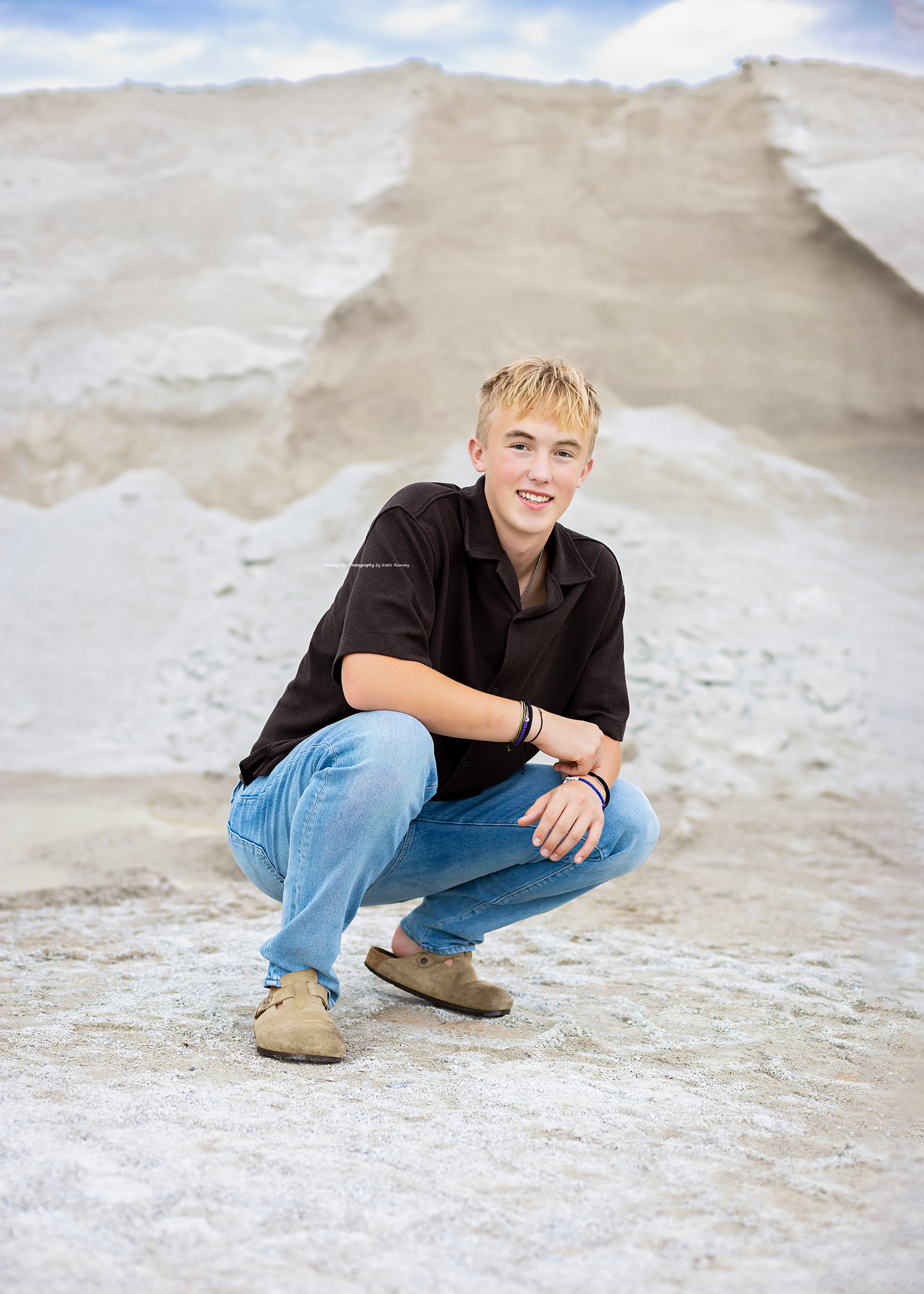 Teenage boy with blonde hair crouching in jeans and black shirt against white sand dunes backdrop.
