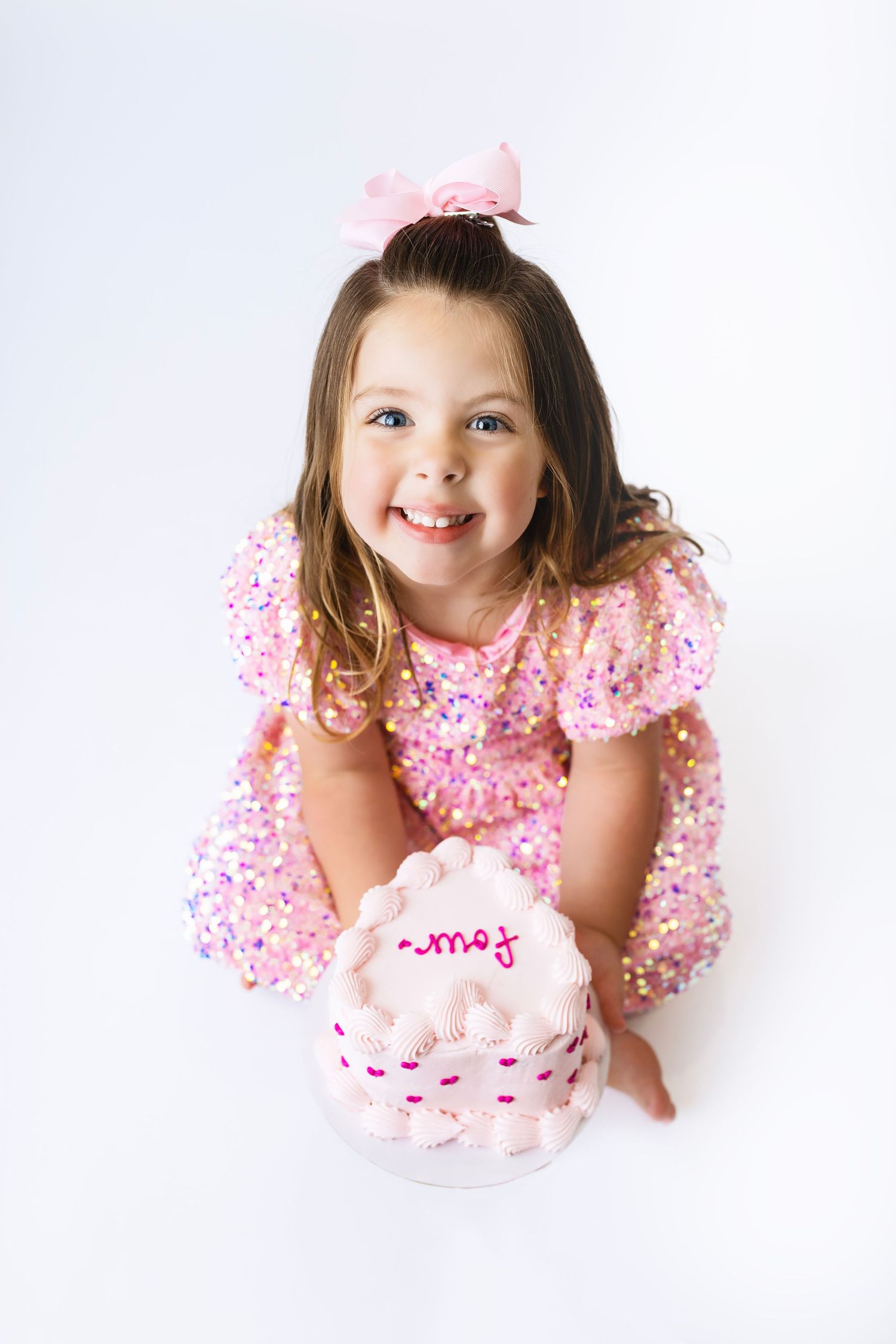Smiling little girl in pink sequin dress with bow sits beside a decorated pink birthday cake on white background.