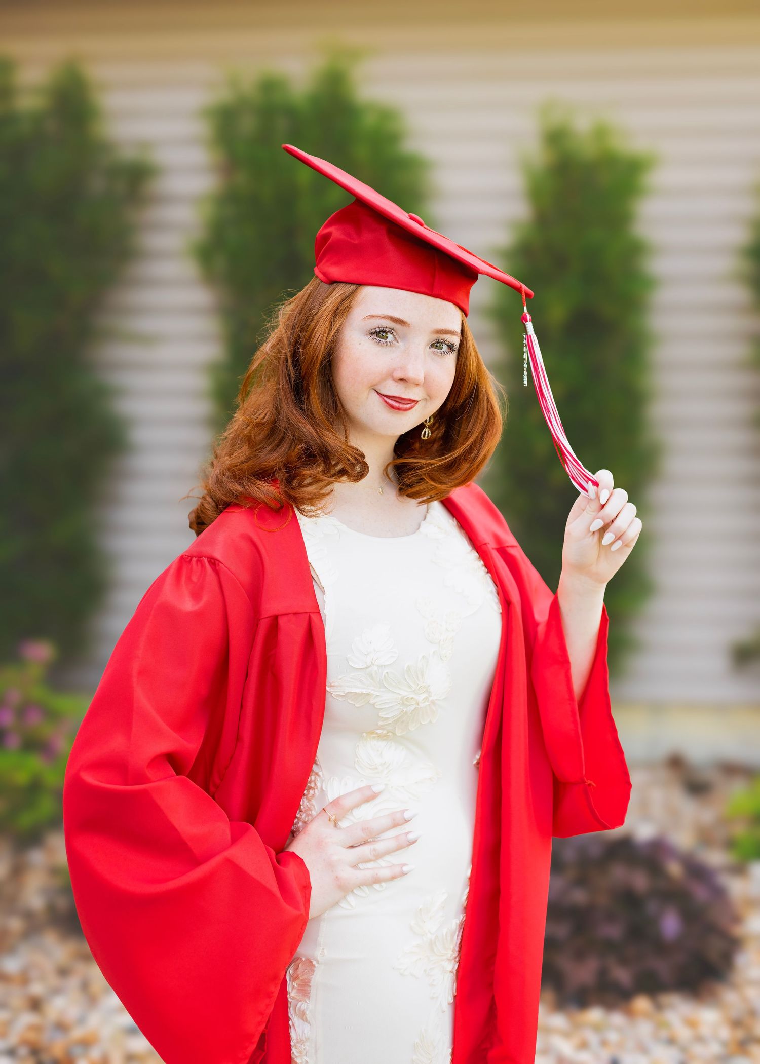 Young woman in red graduation cap and gown holding tassel, posing outdoors in white dress for senior portrait.