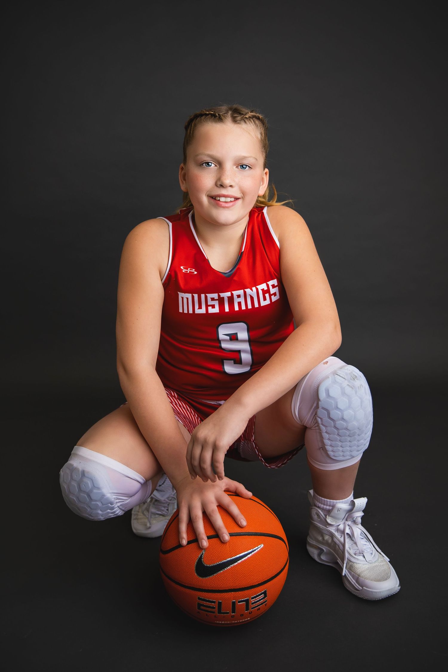 Young girl in Mustangs number 9 red jersey kneels with a Nike Elite basketball.