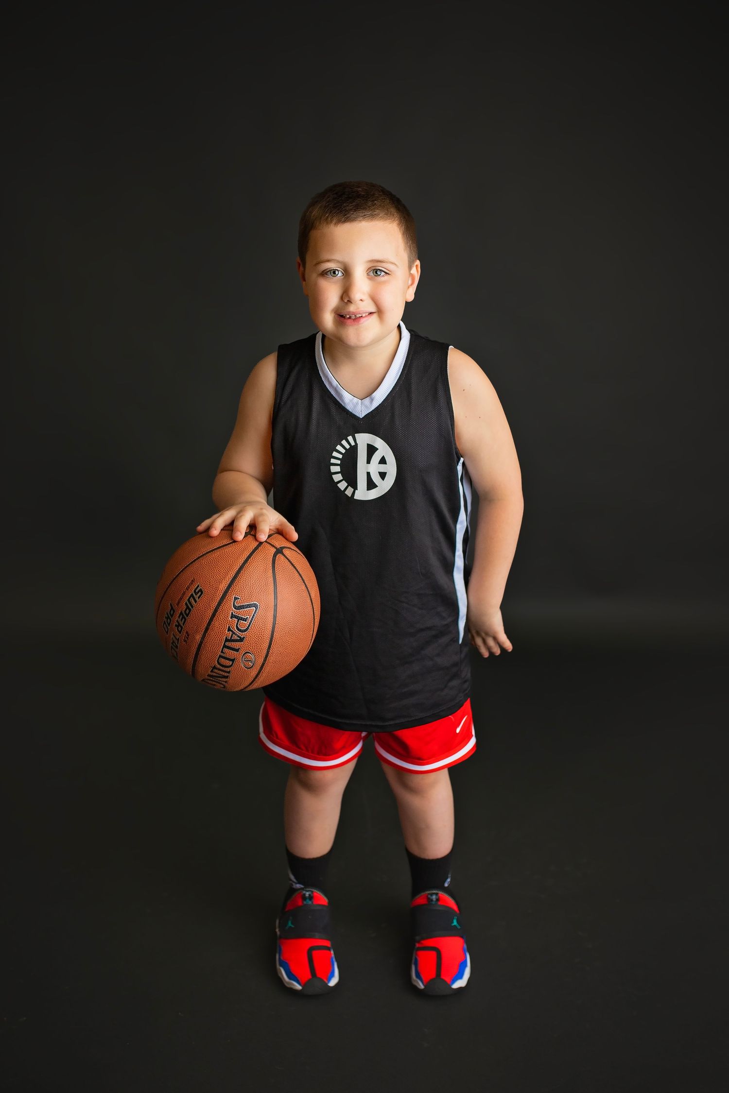 Young boy in Brooklyn Nets jersey holding Spalding basketball standing on dark background.