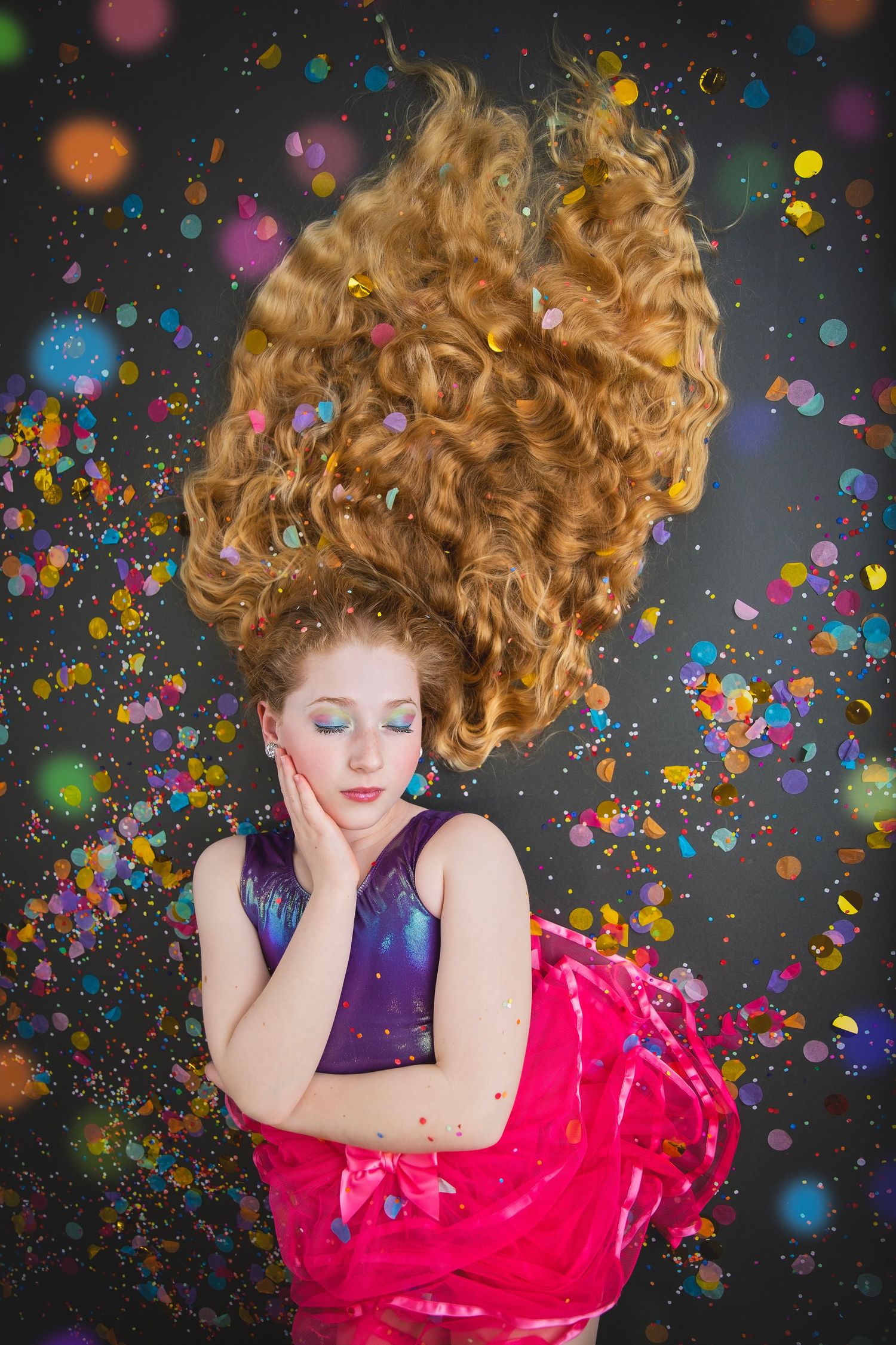 A young girl with long curly red hair lies on a dark floor in a purple top and pink skirt with confetti falling.