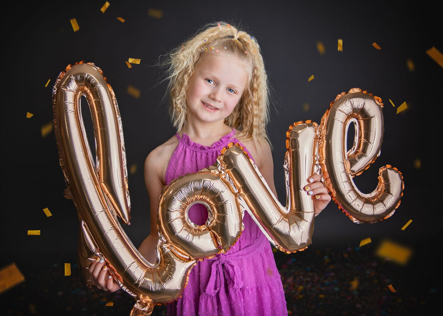 Young girl in pink dress holding gold love balloon with confetti falling on dark background.