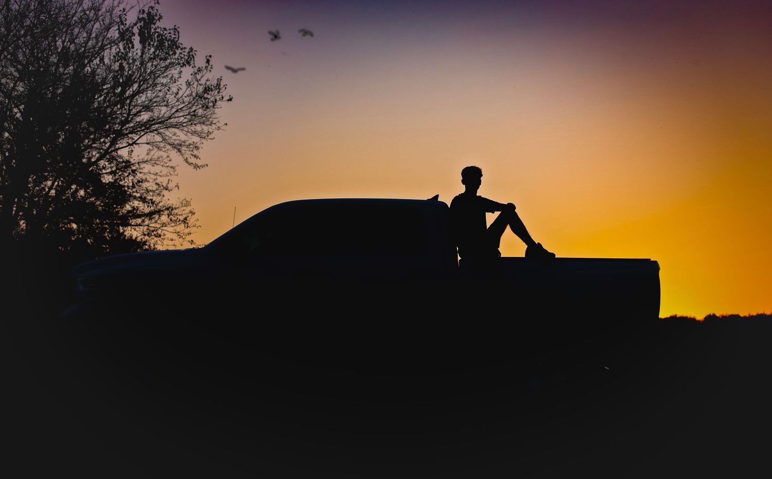 Silhouette of a teen boy sitting on a truck bed against a vibrant orange and purple sunset sky.