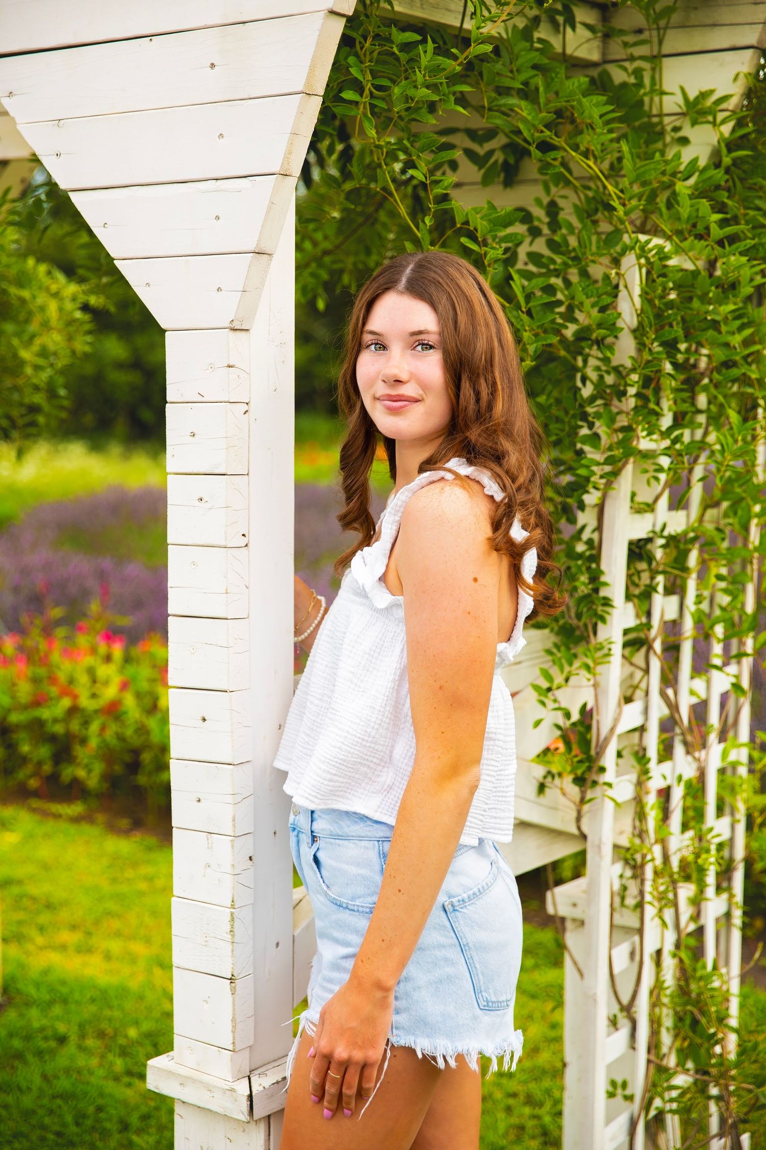 Teen girl in white top and denim shorts posing by a white garden arbor.