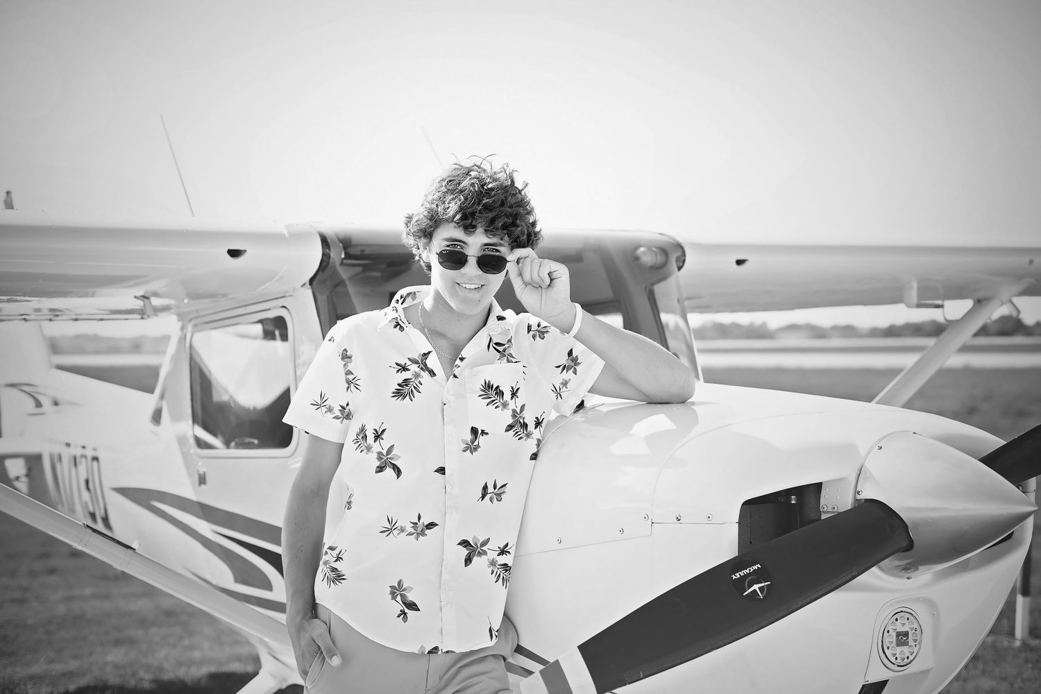 Curly-haired teen boy in floral shirt poses beside a small propeller plane in black and white.