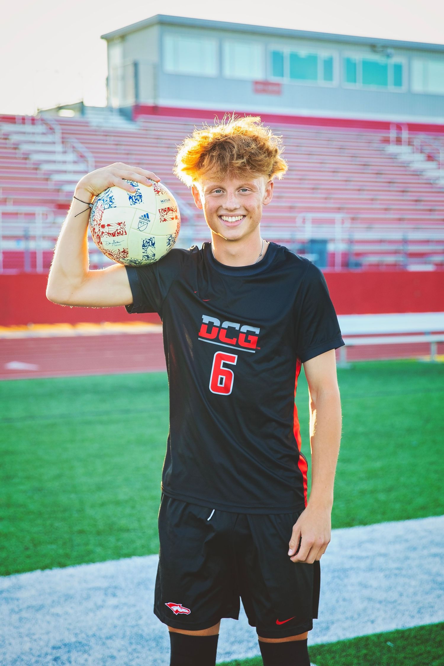 Smiling teen soccer player in DCG number 6 jersey holding ball at outdoor stadium.