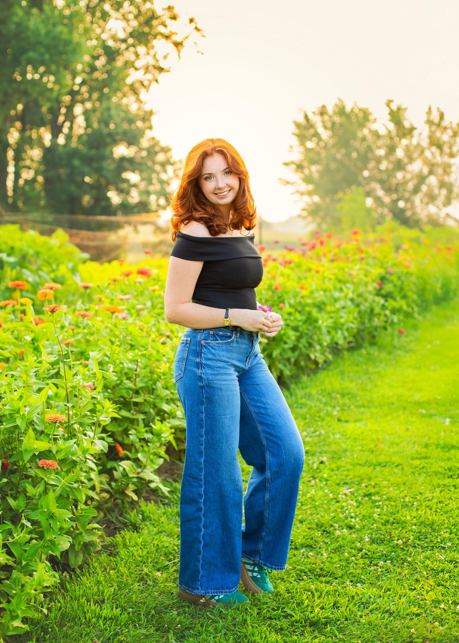 Redhead woman in black top and wide-leg jeans posing in a vibrant flower garden at sunset.