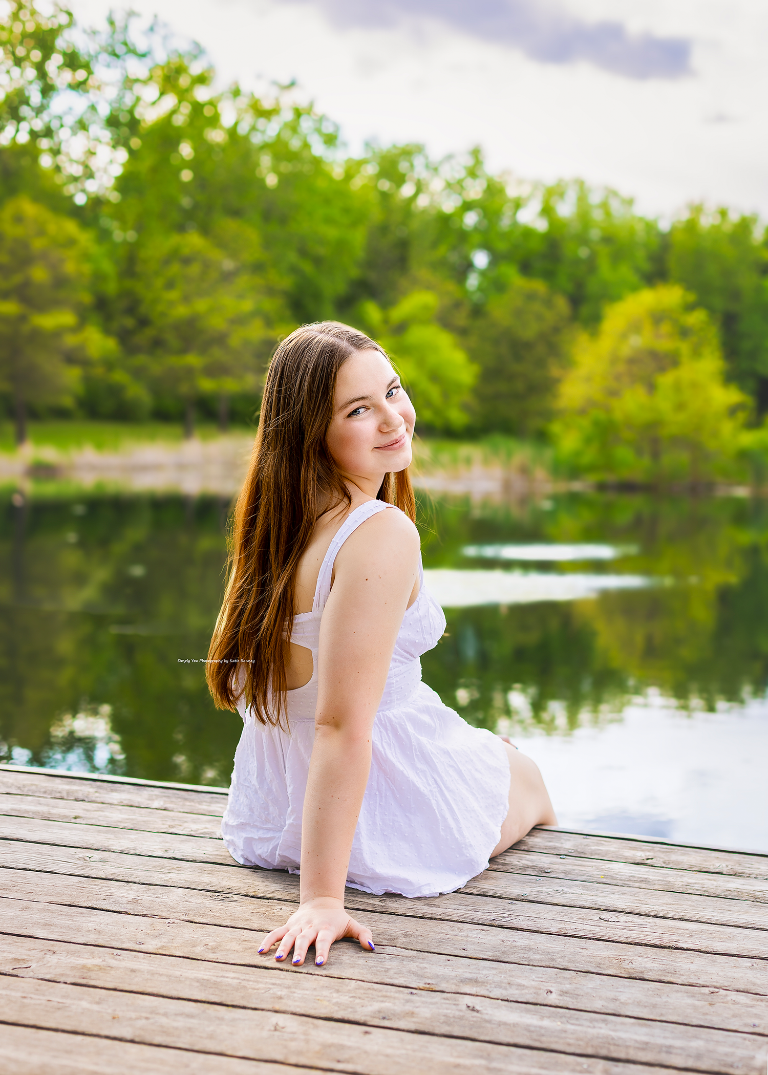 Young girl in white dress sitting on wooden dock by calm lake surrounded by lush green trees on sunny day.