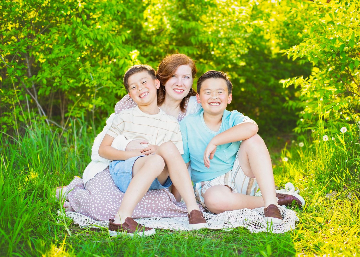 A smiling woman sits on a blanket in green grass with two happy boys beside her.