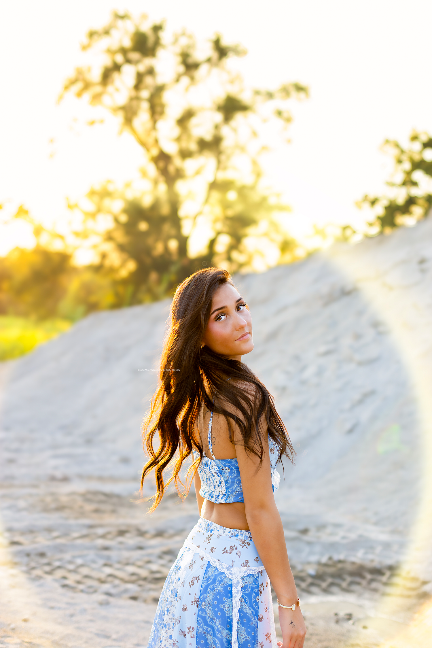 Young woman in blue floral two-piece outfit poses outdoors at golden hour with sandy hill and trees behind her.