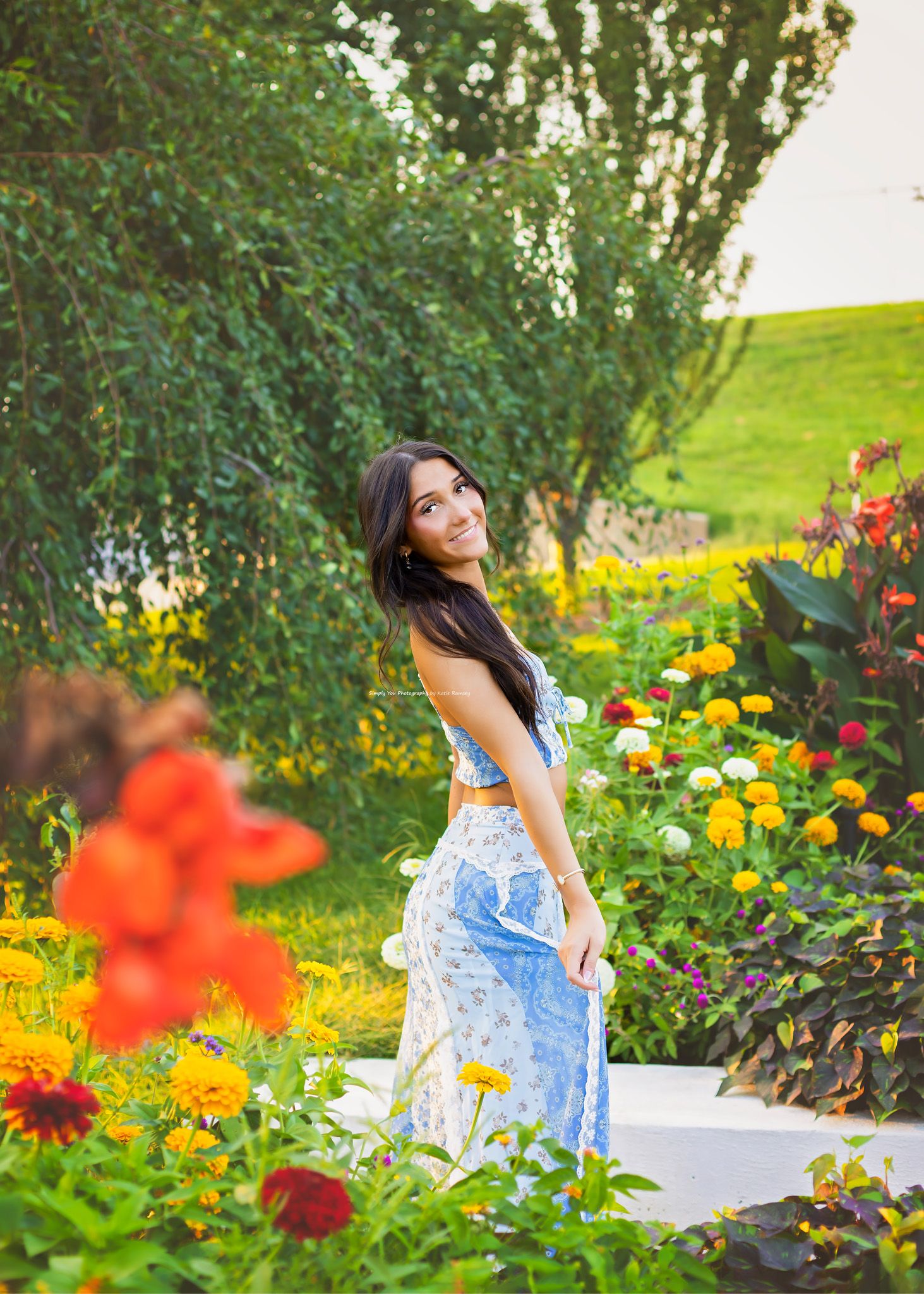 Young woman in blue floral two-piece outfit smiling in a vibrant garden with colorful flowers.