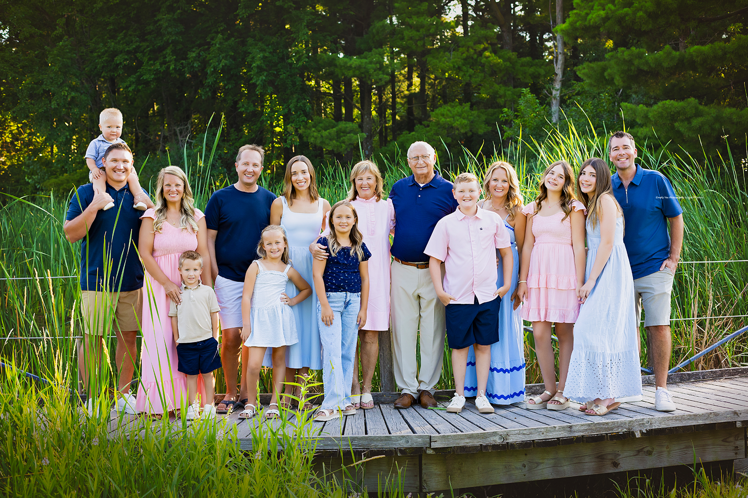 Large family group portrait on wooden dock surrounded by lush green trees, wearing coordinated pastel outfits.