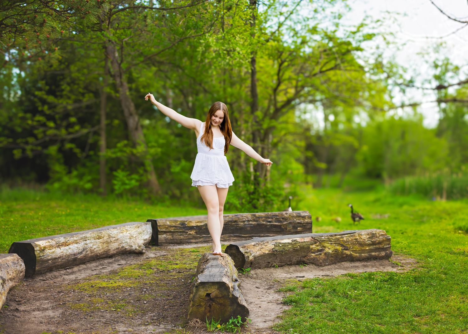Young woman in white romper balancing on a log in a lush green park with arms outstretched.
