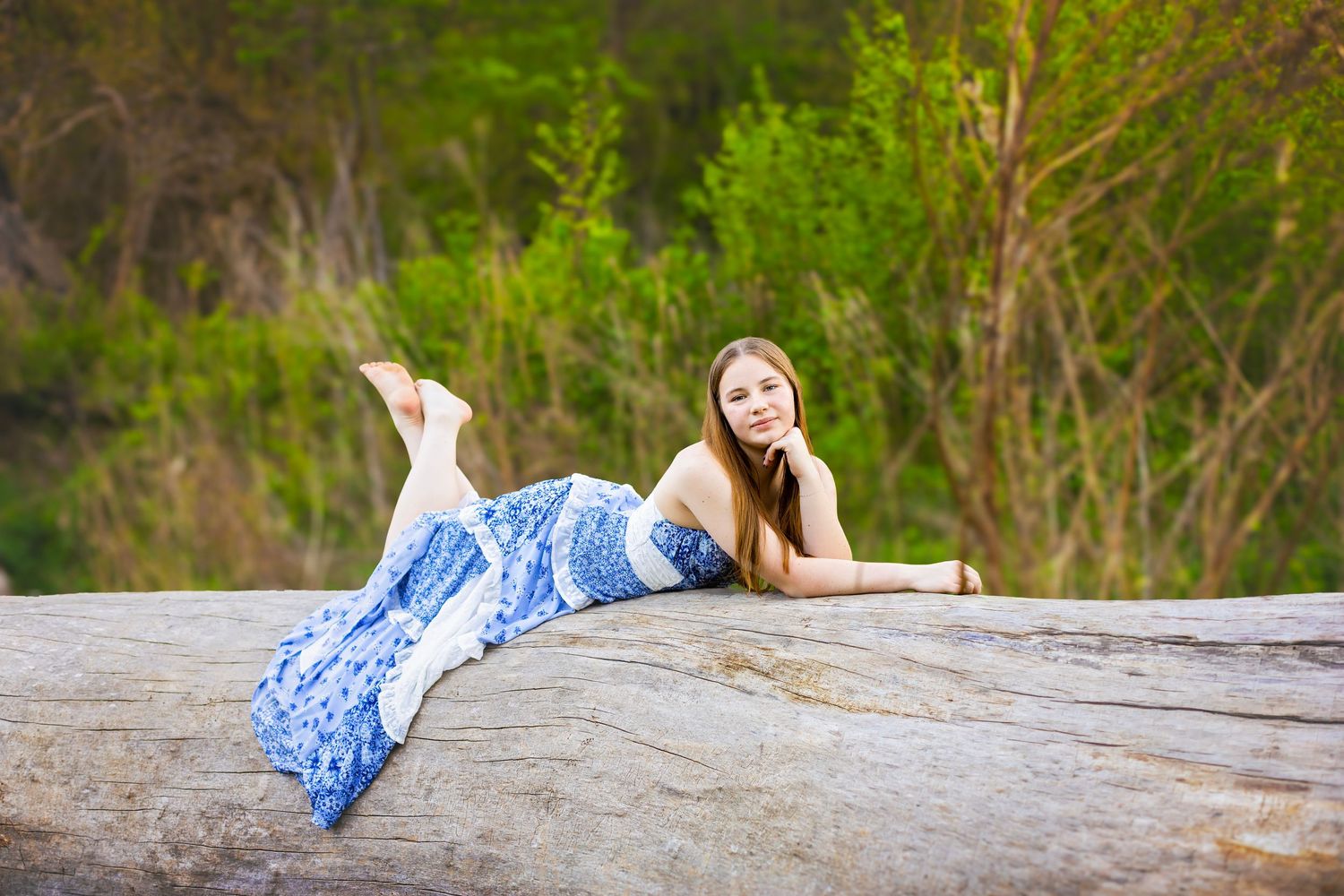 Young woman in blue floral dress lounging on a large rock surrounded by lush green trees outdoors.