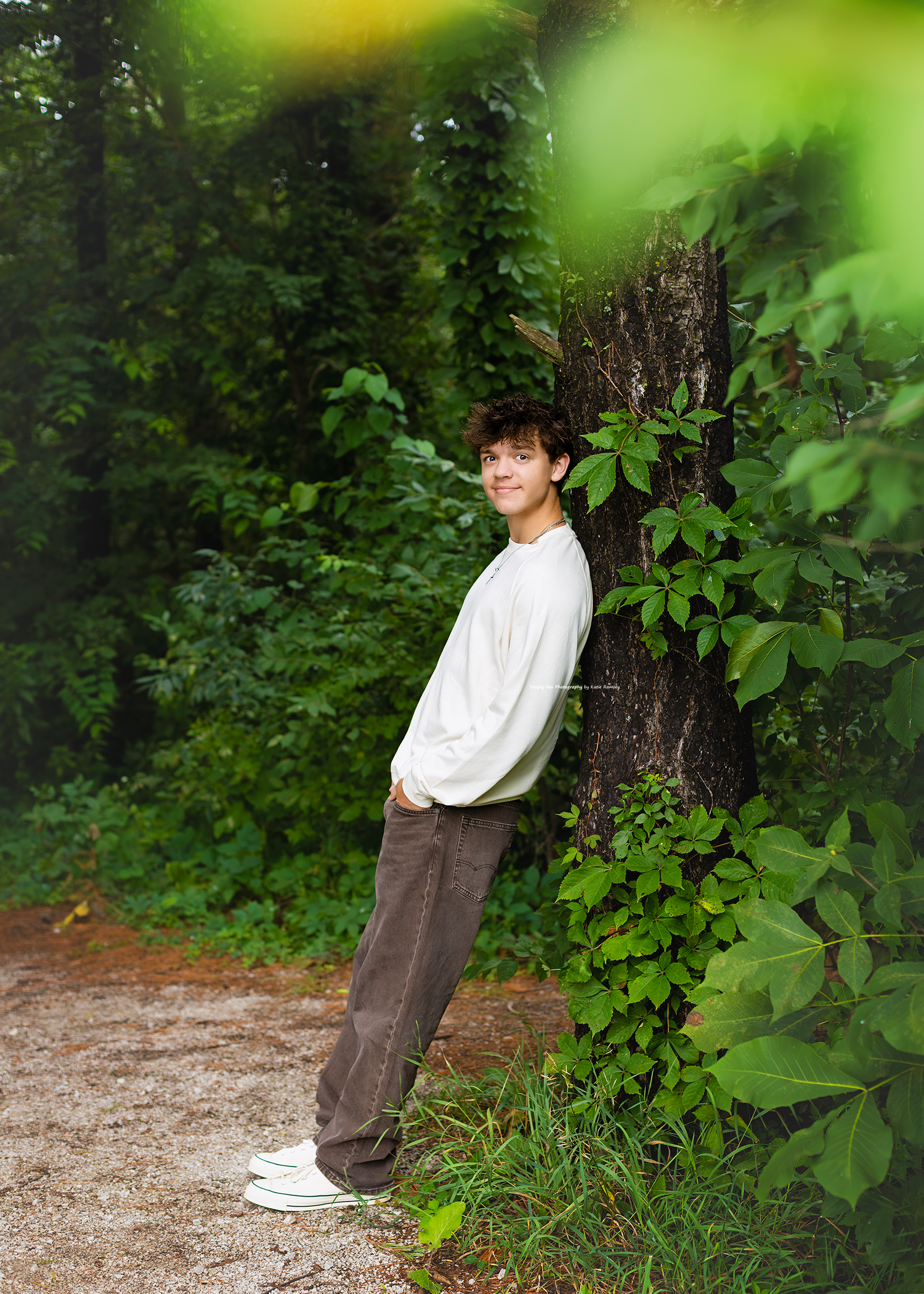 Young man in white sweatshirt and brown pants leaning against a tree in a lush green forest setting.