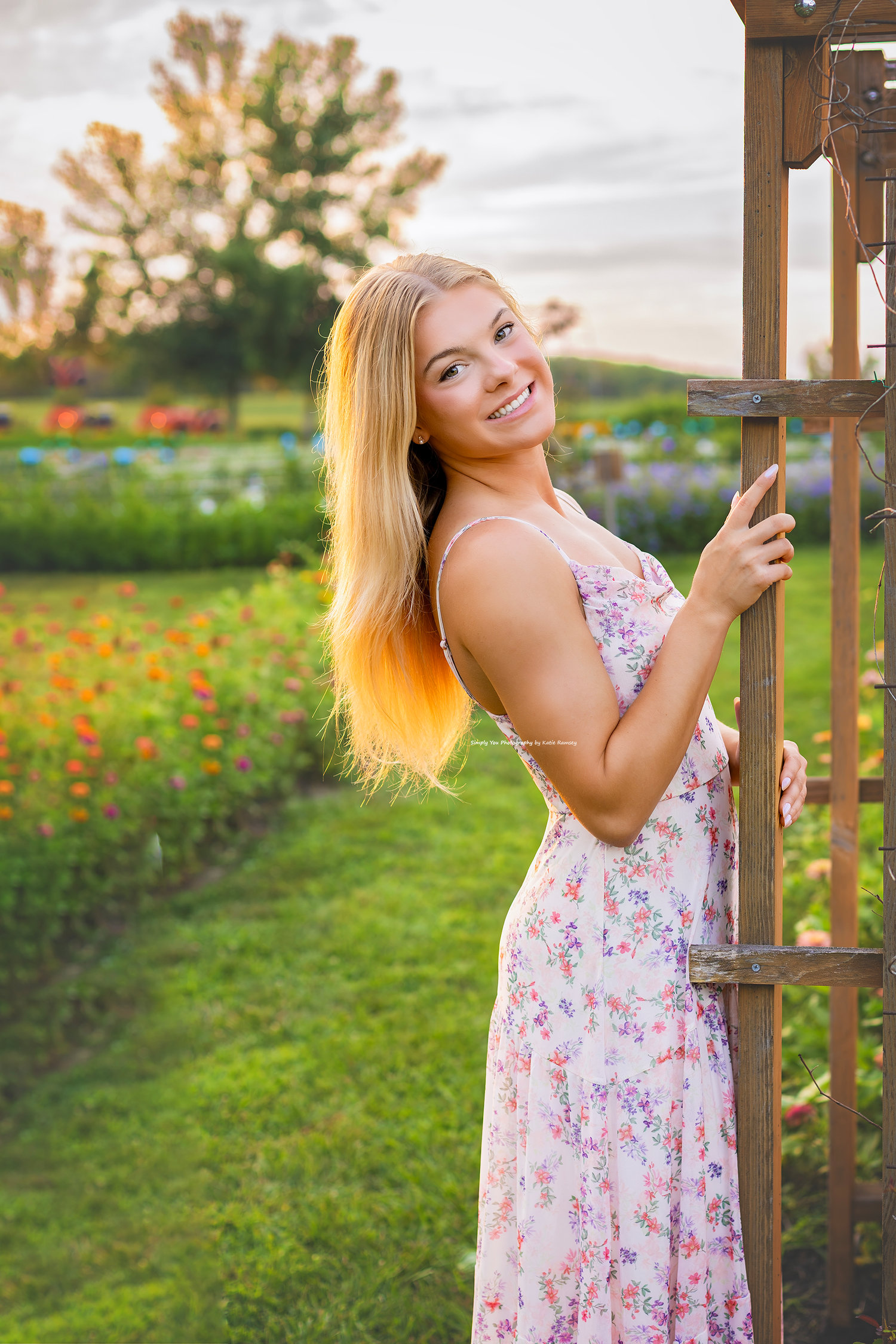 Smiling blonde woman in floral sundress standing by wooden garden trellis with colorful flower beds in background.
