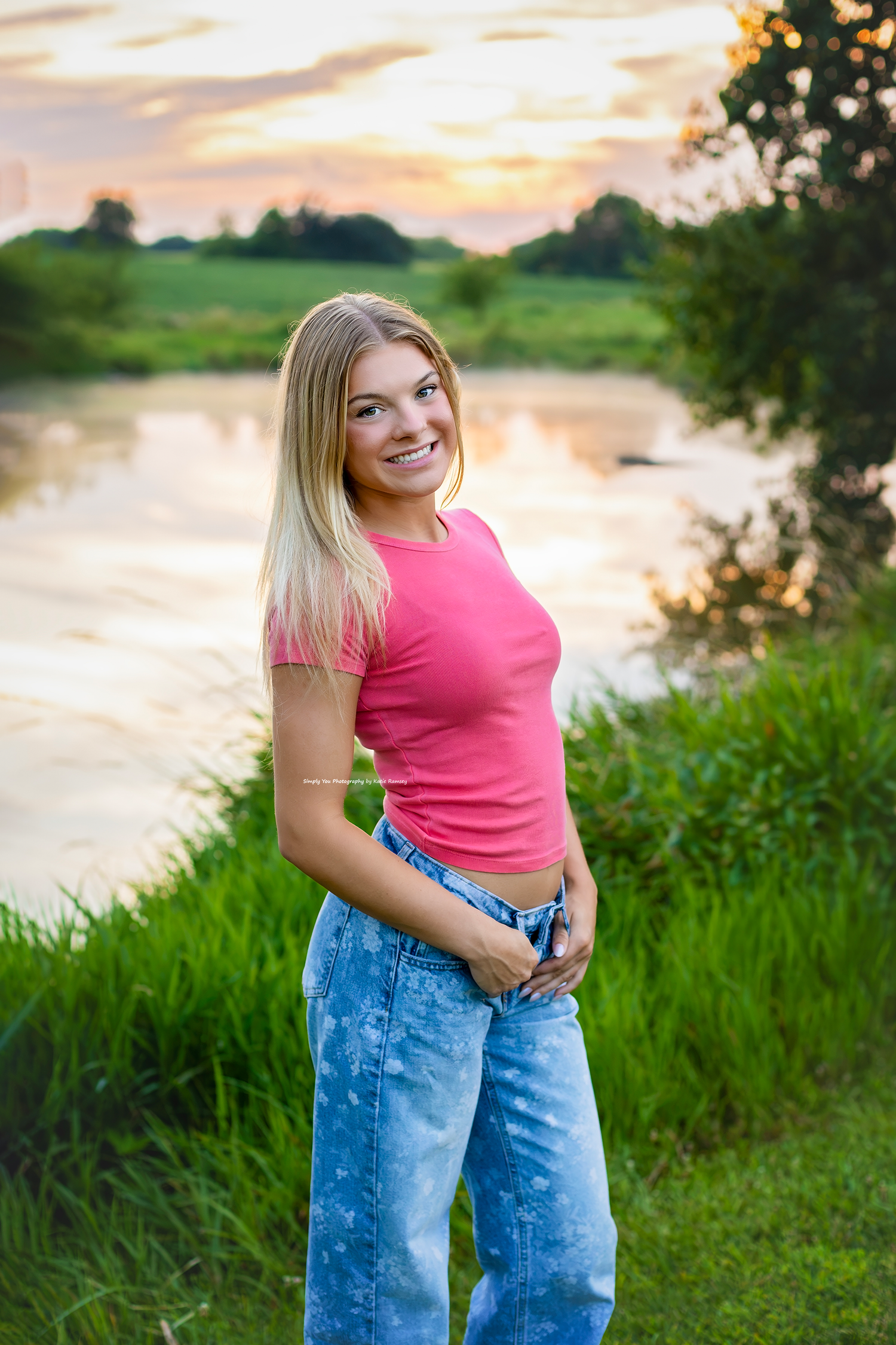 Smiling blonde teen in pink top and jeans poses by a serene river at sunset with lush green grass.