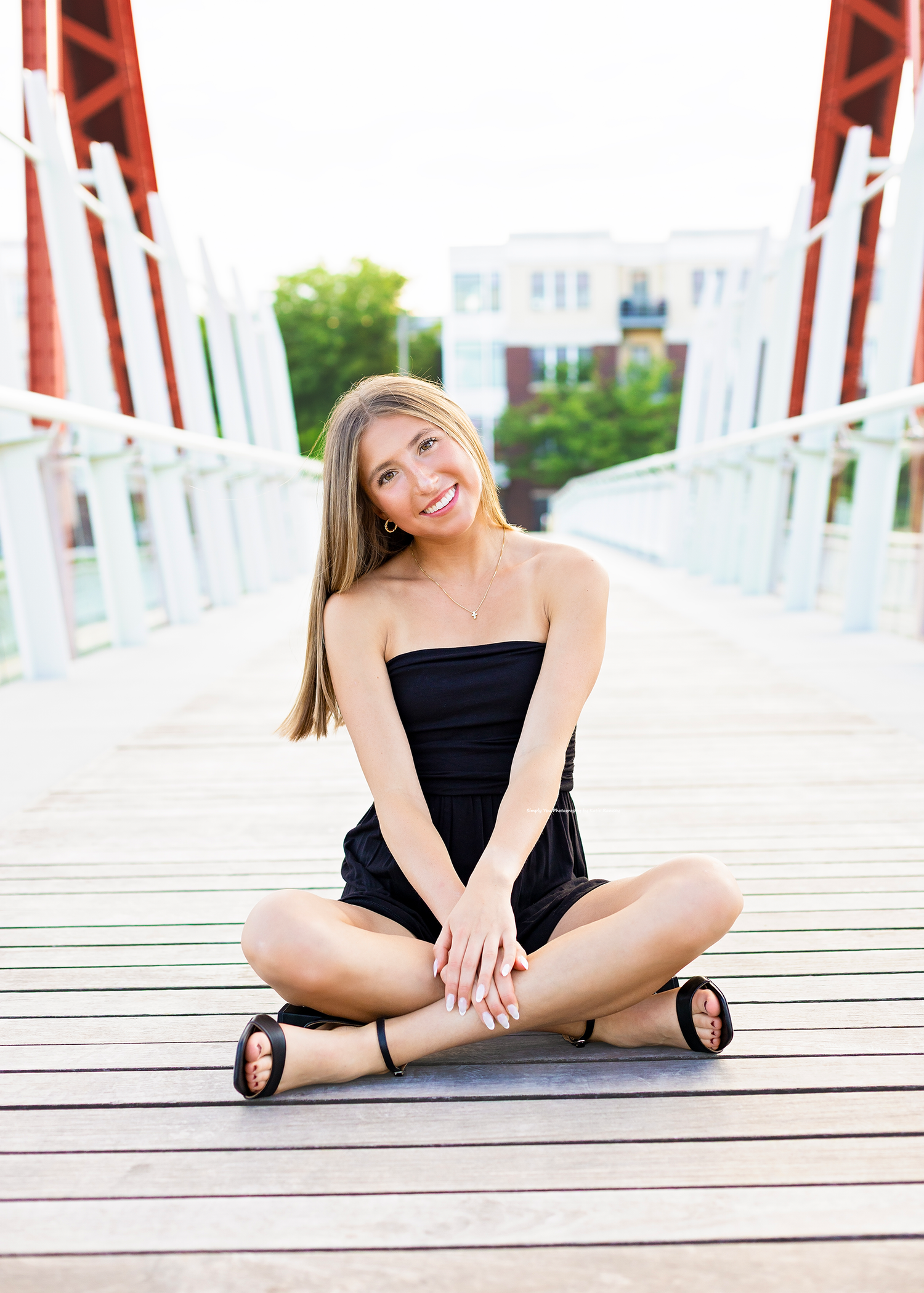Smiling young woman in black romper sitting cross-legged on wooden bridge outdoors.