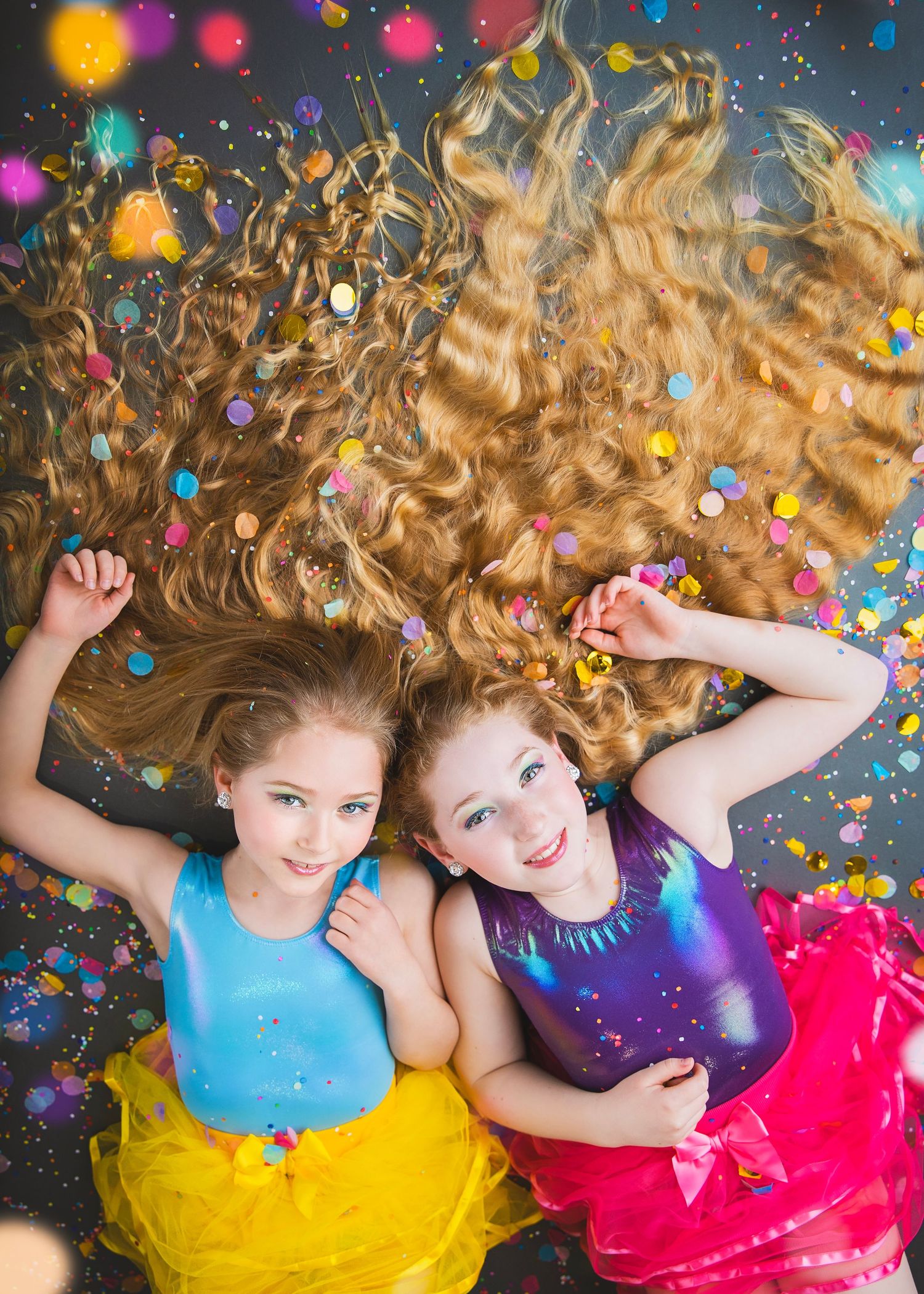 Two girls in colorful tutus lying on floor with confetti and long wavy hair spread around them.