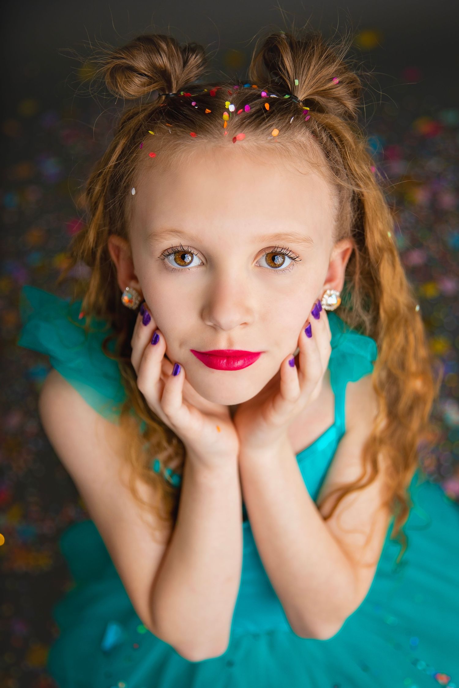 Young girl in teal dress with purple nails posing among colorful confetti.