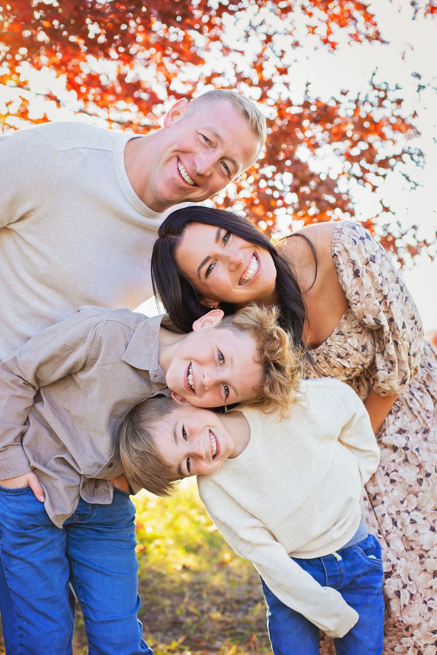 A happy family of four laughs together outdoors beneath vibrant red autumn foliage in a warm fall portrait.