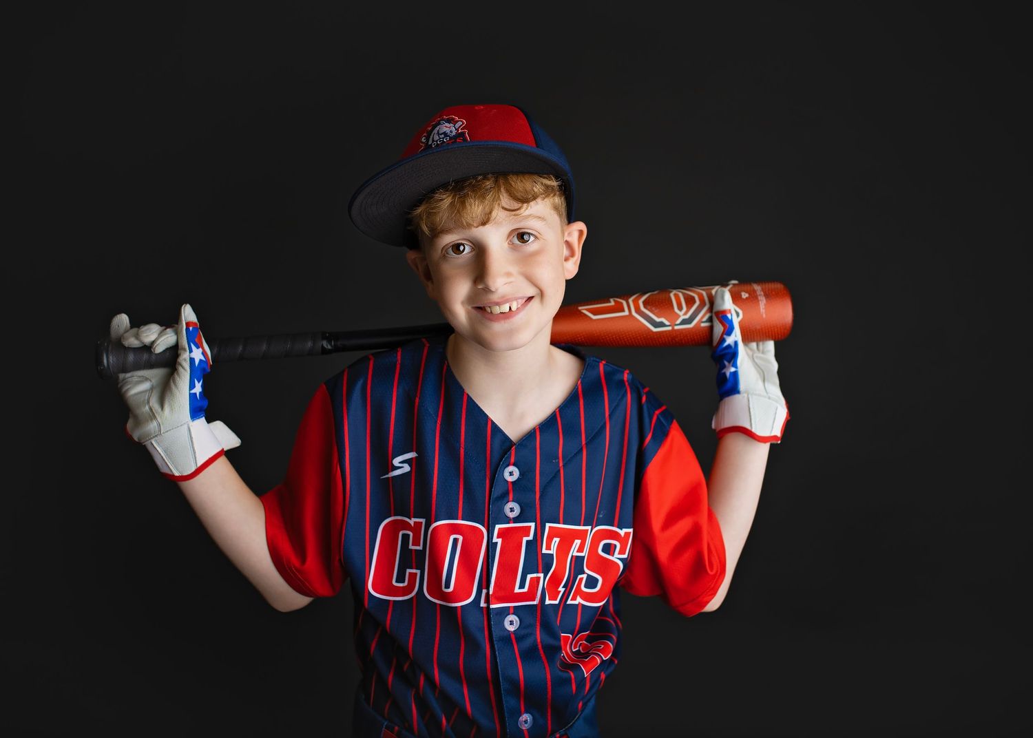 Young boy in Colts baseball uniform smiling with bat over shoulder on dark background.