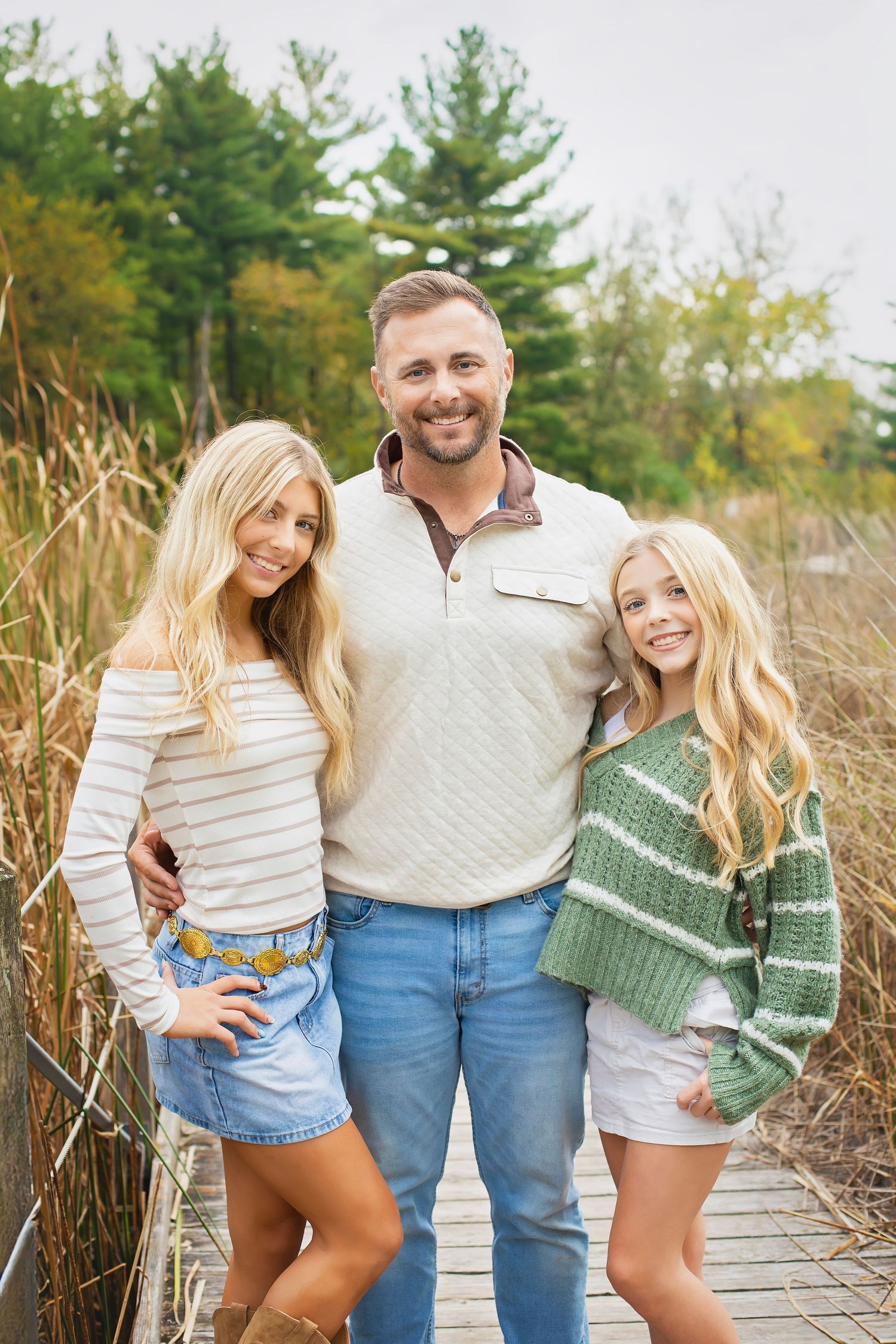 A smiling family of three, father, mother, and daughter, pose together on a boardwalk amid tall golden reeds.