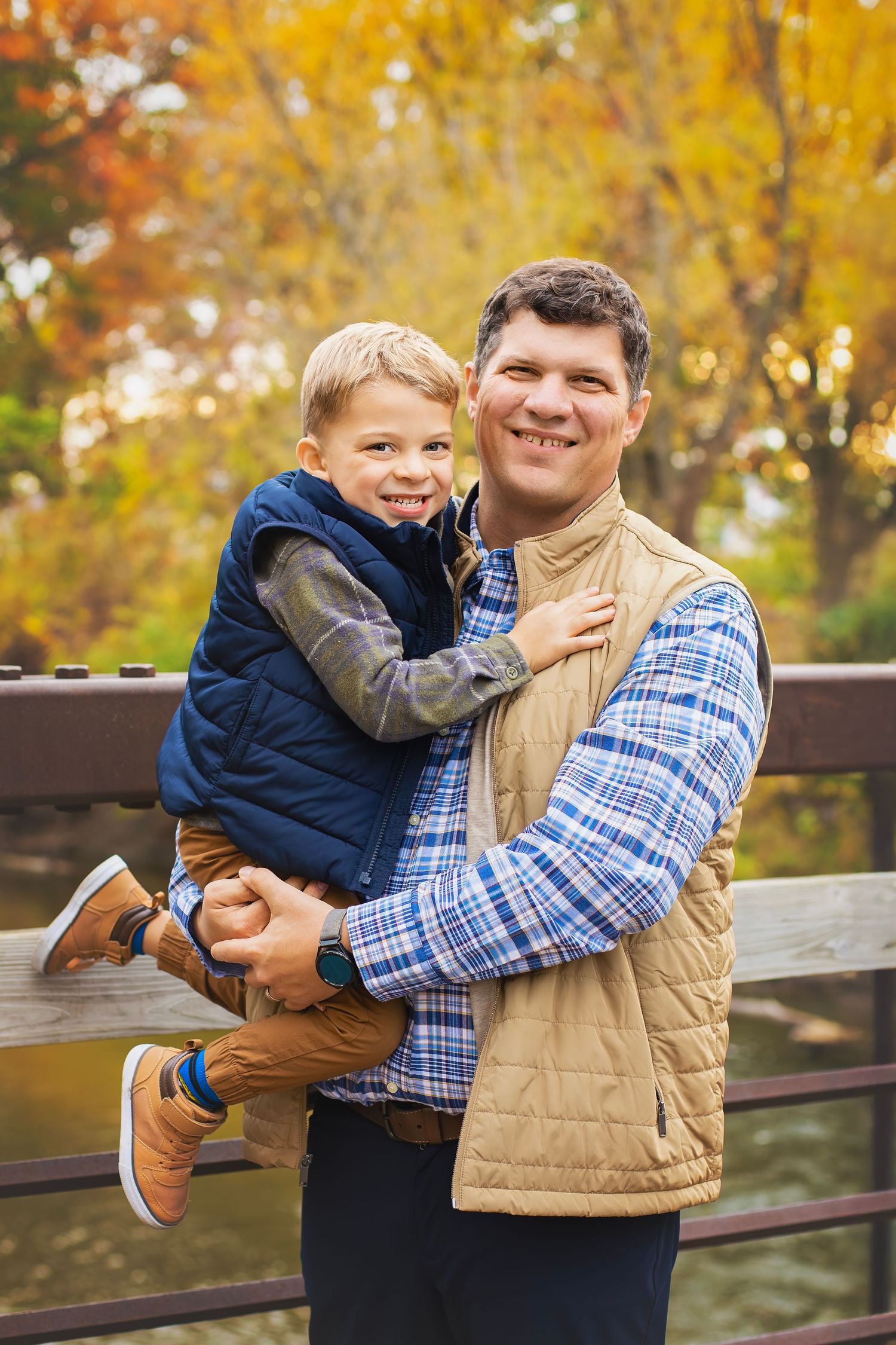 A smiling father holds his young son while standing on a bridge surrounded by colorful fall foliage.