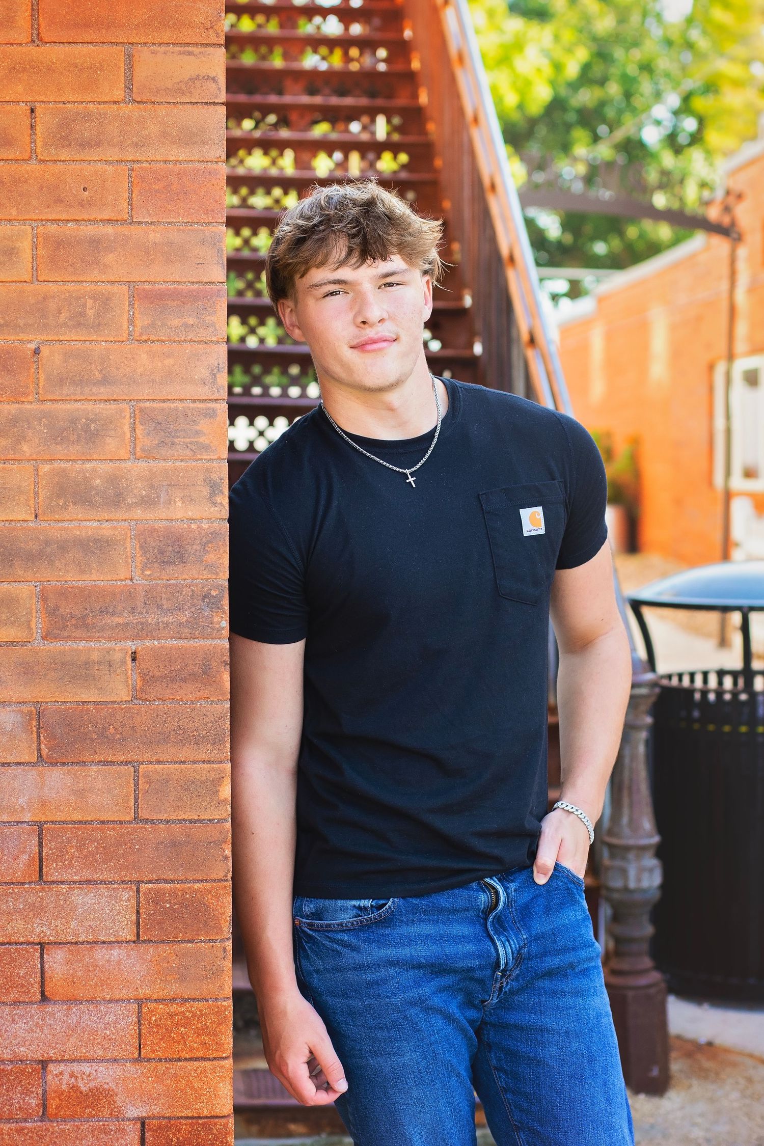 Teenage boy in black Carhartt shirt leans against red brick wall in outdoor setting.