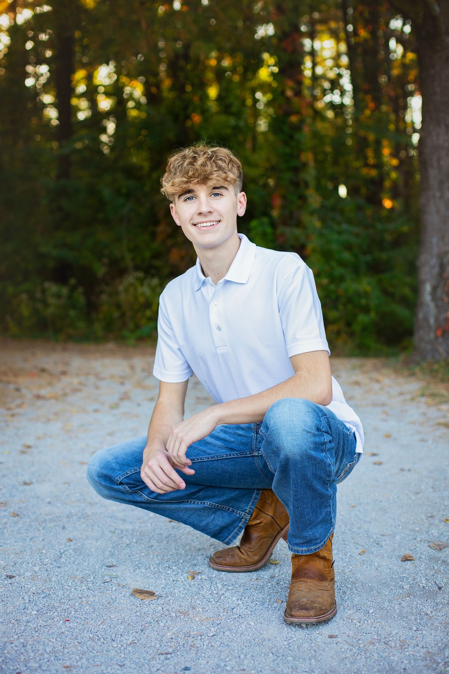 Smiling curly-haired teen boy crouching in white polo and jeans on a gravel path in autumn woods.