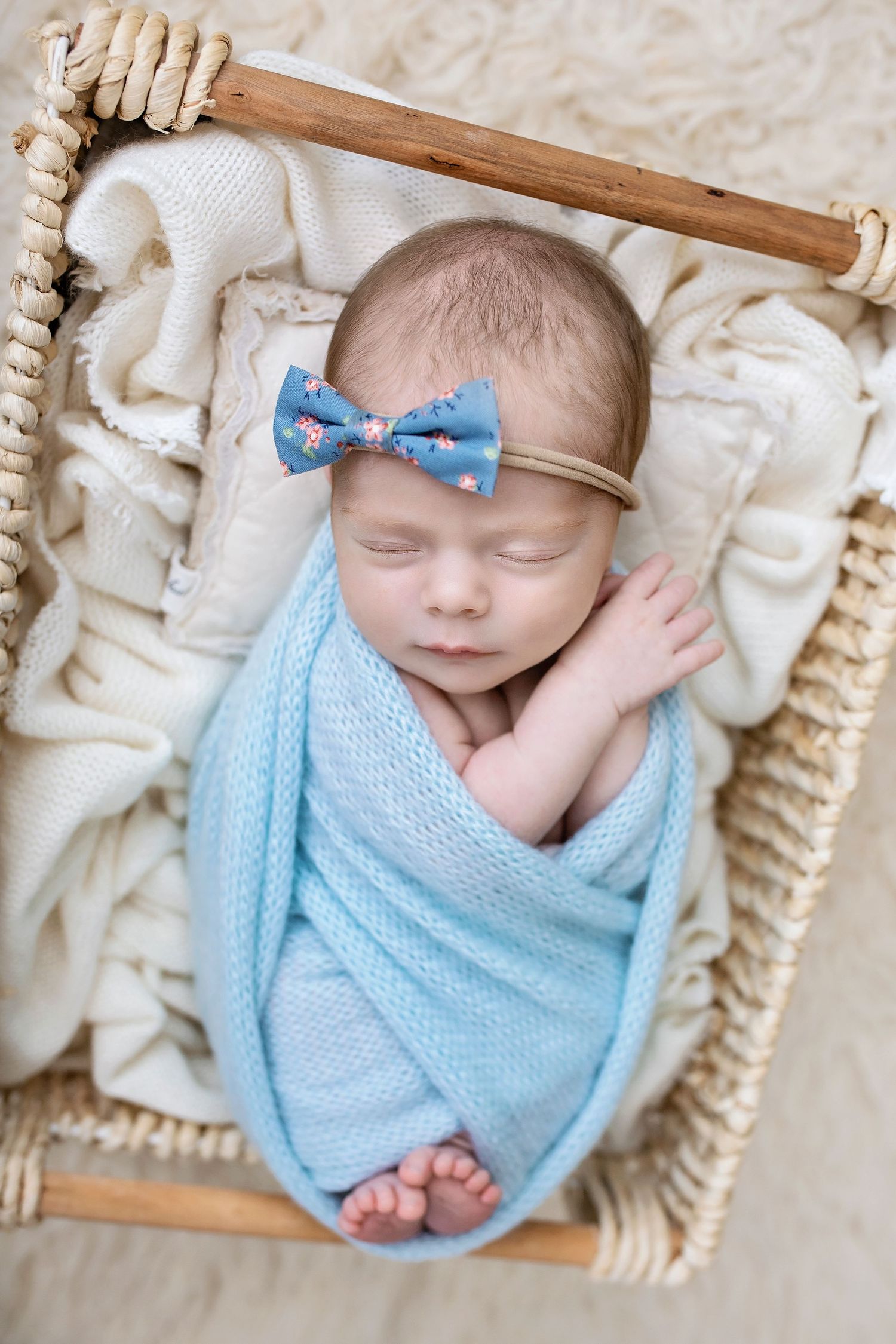 Sleeping newborn baby girl swaddled in blue knit wrap with floral bow headband in wicker basket.
