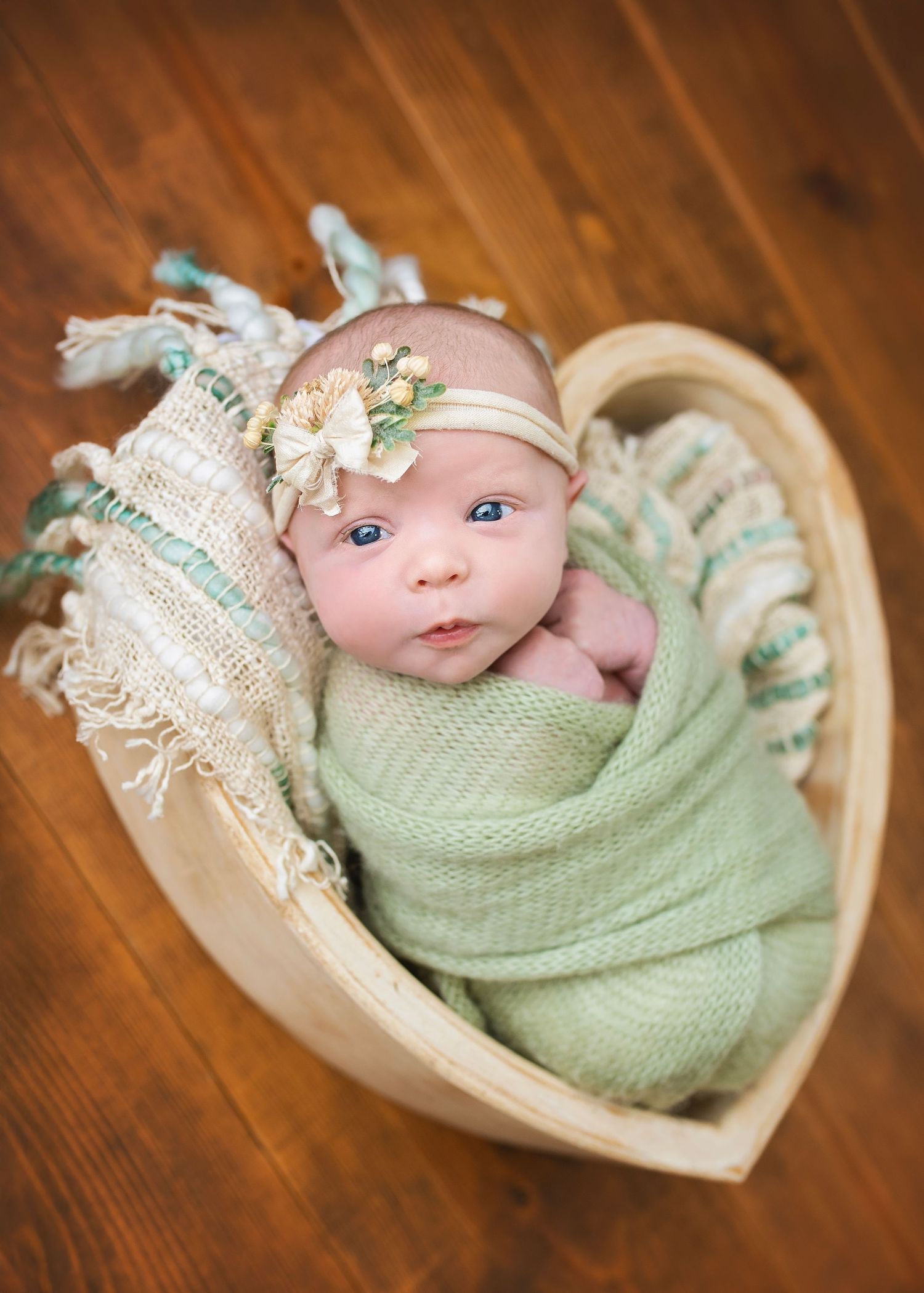 Newborn baby girl swaddled in green wrap with floral headband posed in heart-shaped wooden bowl.
