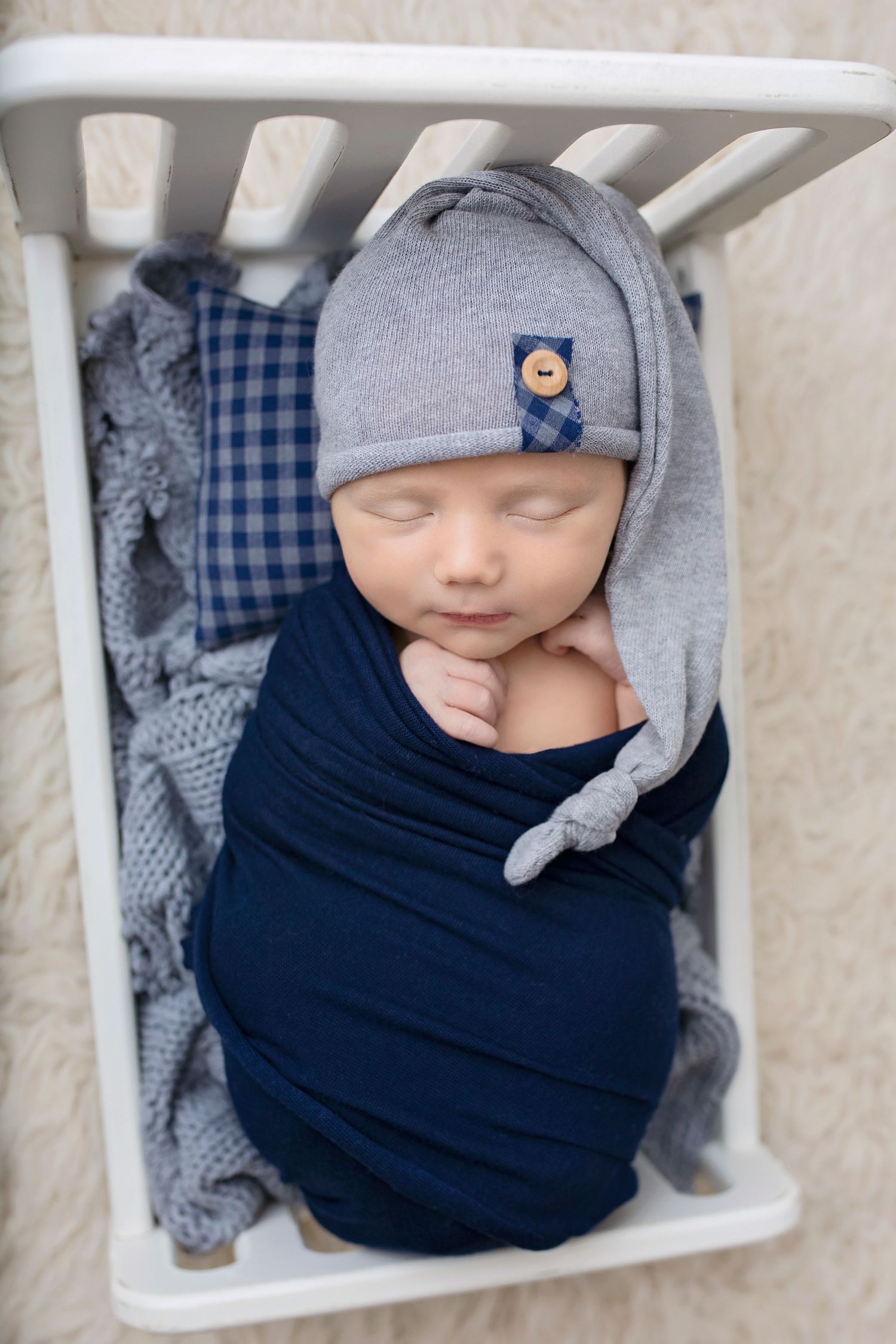 Sleeping newborn baby swaddled in navy wrap wearing a gray beanie hat in a white mini crib.