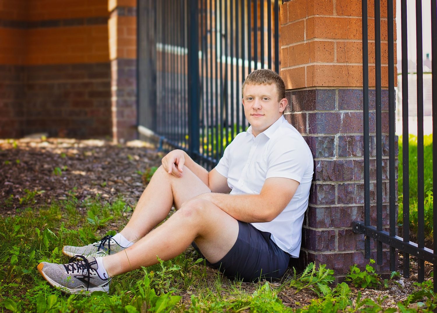 Young male athlete in white polo and shorts sitting relaxed against brick wall outdoors.
