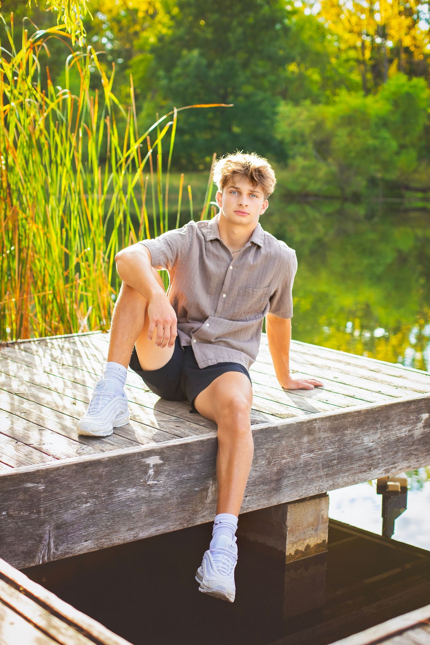 Young man in gray shirt and black shorts sits on the edge of a wooden dock near reeds and a calm lake.