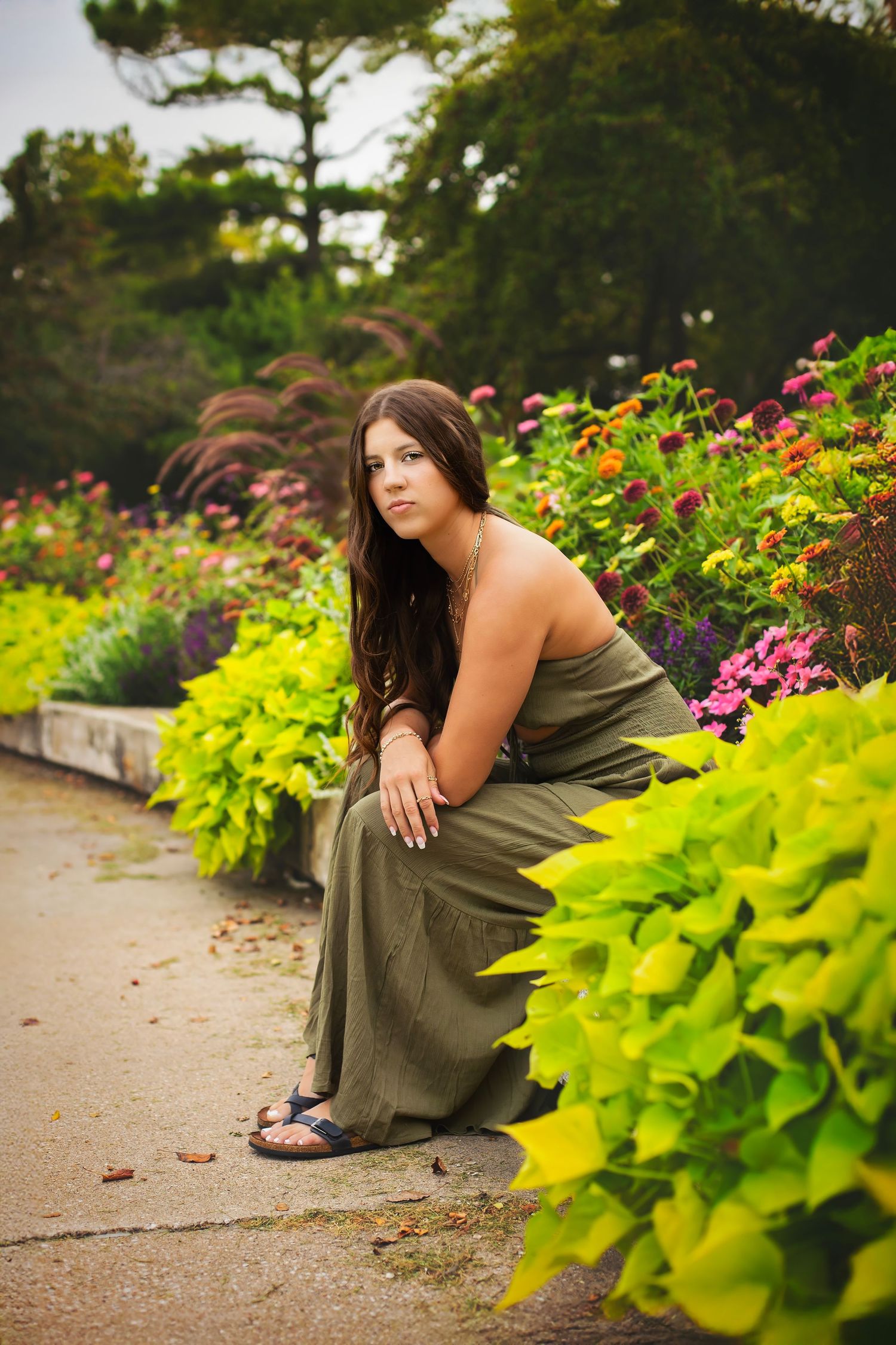 Young woman in olive green maxi dress sits along a vibrant garden path surrounded by flowers.